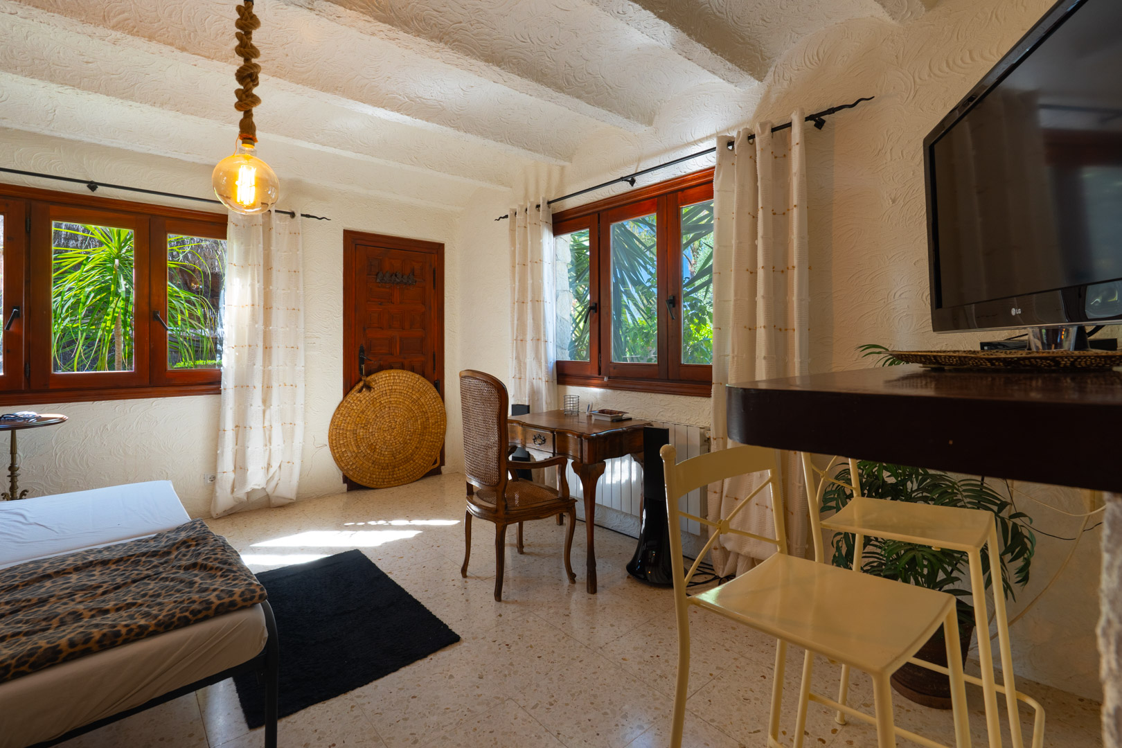 Rustikales Gästezimmer mit weißen Balkendecken, klassischem Holzschreibtisch und dunklen Fensterrahmen mit Blick auf das mediterrane Grün.