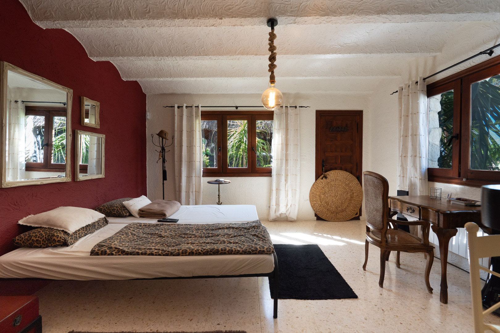 Rustikales Schlafzimmer mit bordeauxroter Akzentwand, antikem Holzschreibtisch und strukturierten Decken. Mit Terrazzoboden und einer markanten Seil-Hängeleuchte.