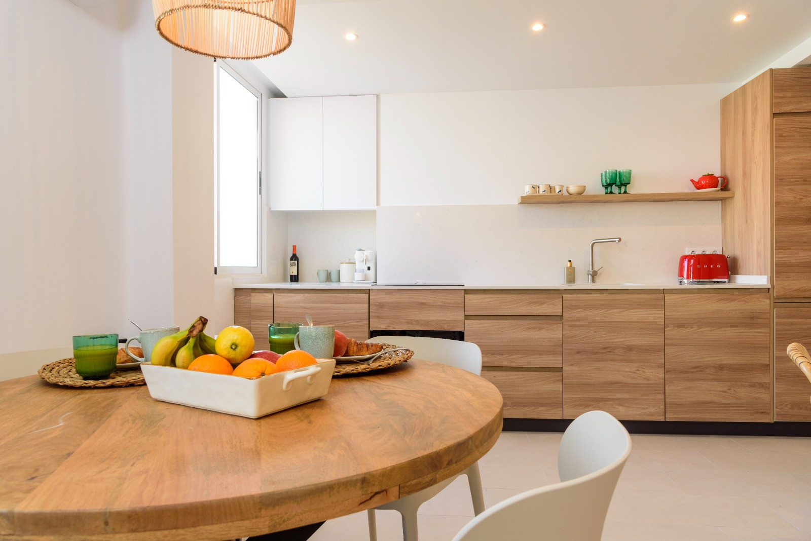 Essbereich mit rundem Holztisch und integrierter Küche mit Schränken in Eichenoptik, weißer Arbeitsplatte und moderner Einbaubeleuchtung.