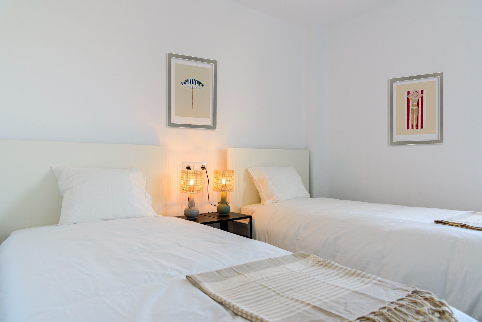 Helles Schlafzimmer mit zwei Einzelbetten, weißen Kopfteilen und Lampen aus Naturfasern. Die weißen Wände sind mit gerahmten Bildern im Küstenstil dekoriert.