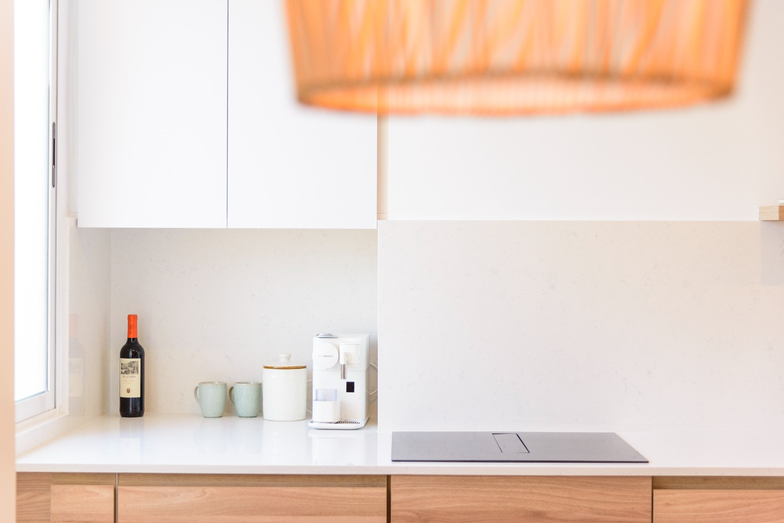 Moderne Küche mit weißer Steinarbeitsplatte, integriertem Induktionskochfeld und Holzunterschränken. Natürliches Licht betont die minimalistische Kaffeeecke und das helle Design.