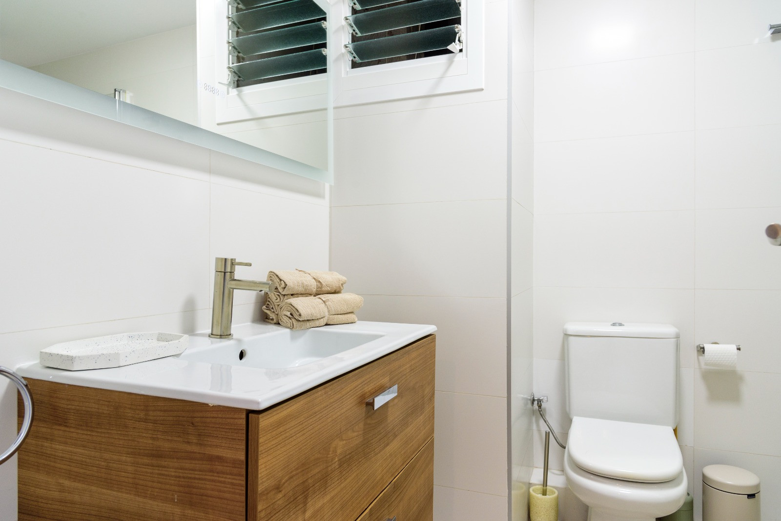 Modernes Badezimmer mit Holzwaschtisch, integriertem Waschbecken und weißen Fliesen. Ausgestattet mit zeitgemäßen Sanitäranlagen und einem Lamellenfenster.