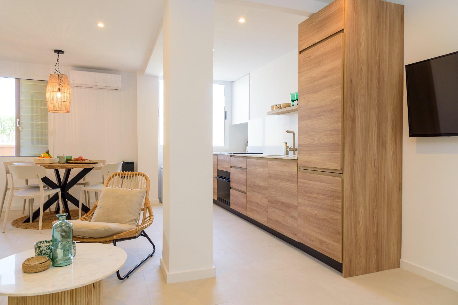Offener Wohnbereich mit moderner Holzküche, Essplatz mit Rattan-Pendelleuchte und hellen Fliesenböden in zeitgemäßem, lichtdurchflutetem Design.