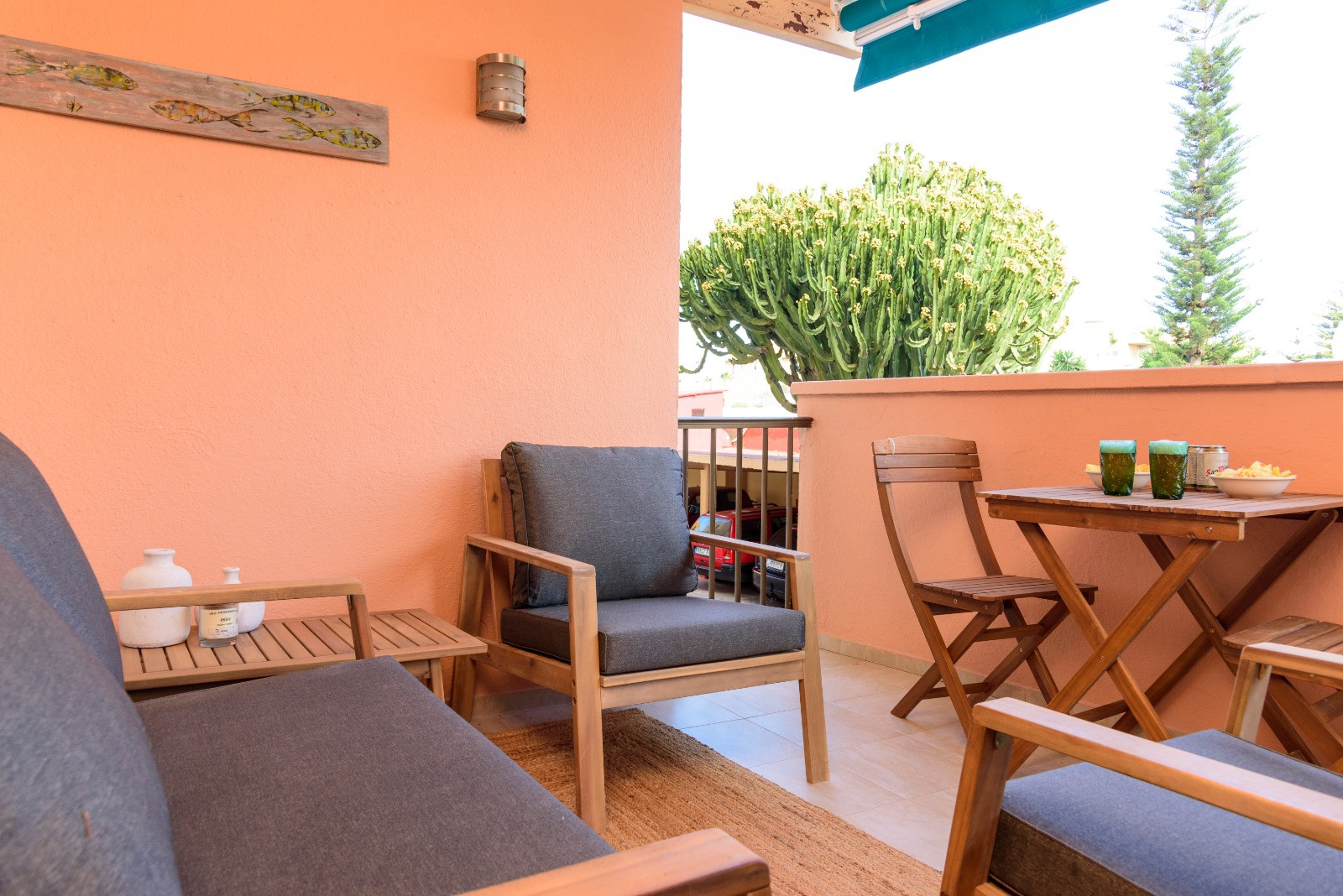 Private Terrasse mit Holzmöbeln, grauen Polstern und Essbereich im Freien, umgeben von mediterraner Vegetation und geschützt durch eine blaue Markise.