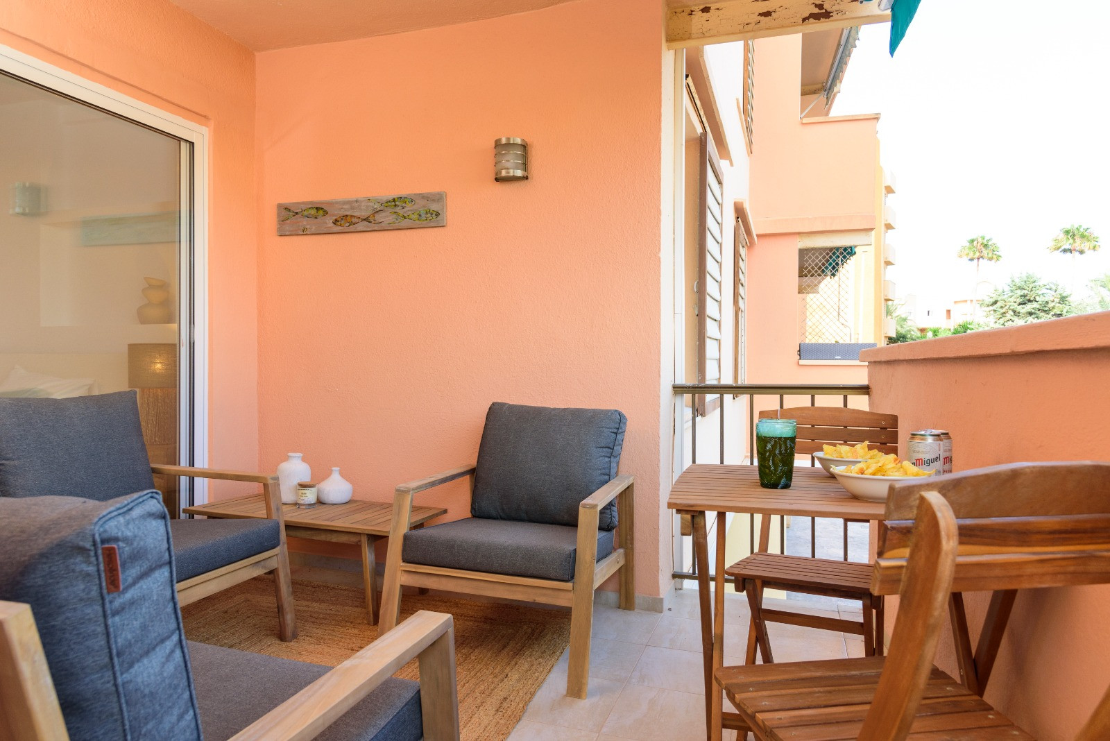 Private Terrasse mit lachsfarbenen Wänden, Holzmöbeln mit grauen Kissen und einem Essbereich im Freien. Große Glasschiebetüren verbinden den Außenbereich mit dem Innenraum.