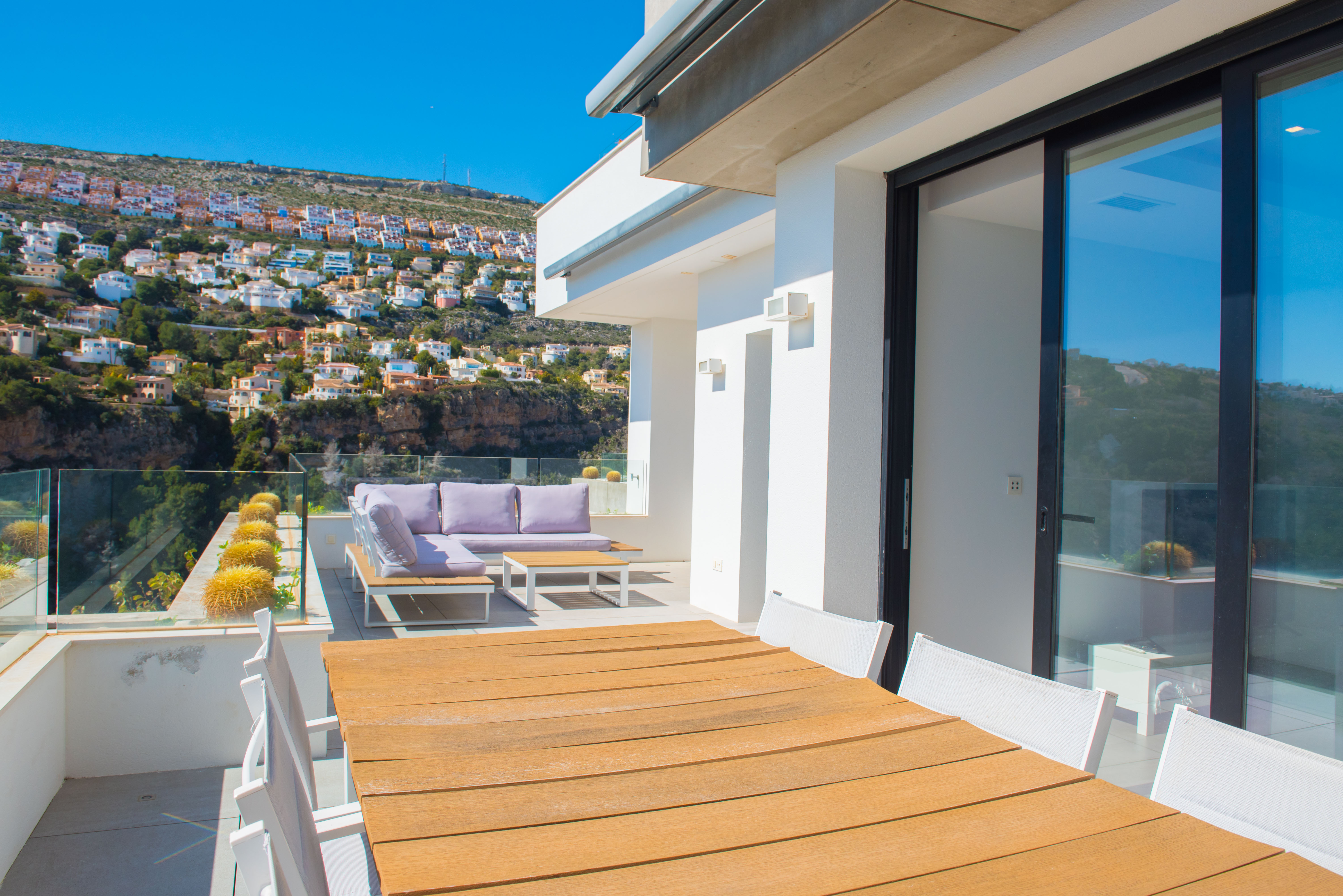 Außen-Terrasse mit Holzesstisch, lila Lounge-Möbeln und Glasgeländern mit Blick auf die bebaute Hügellandschaft von Benitachell.
