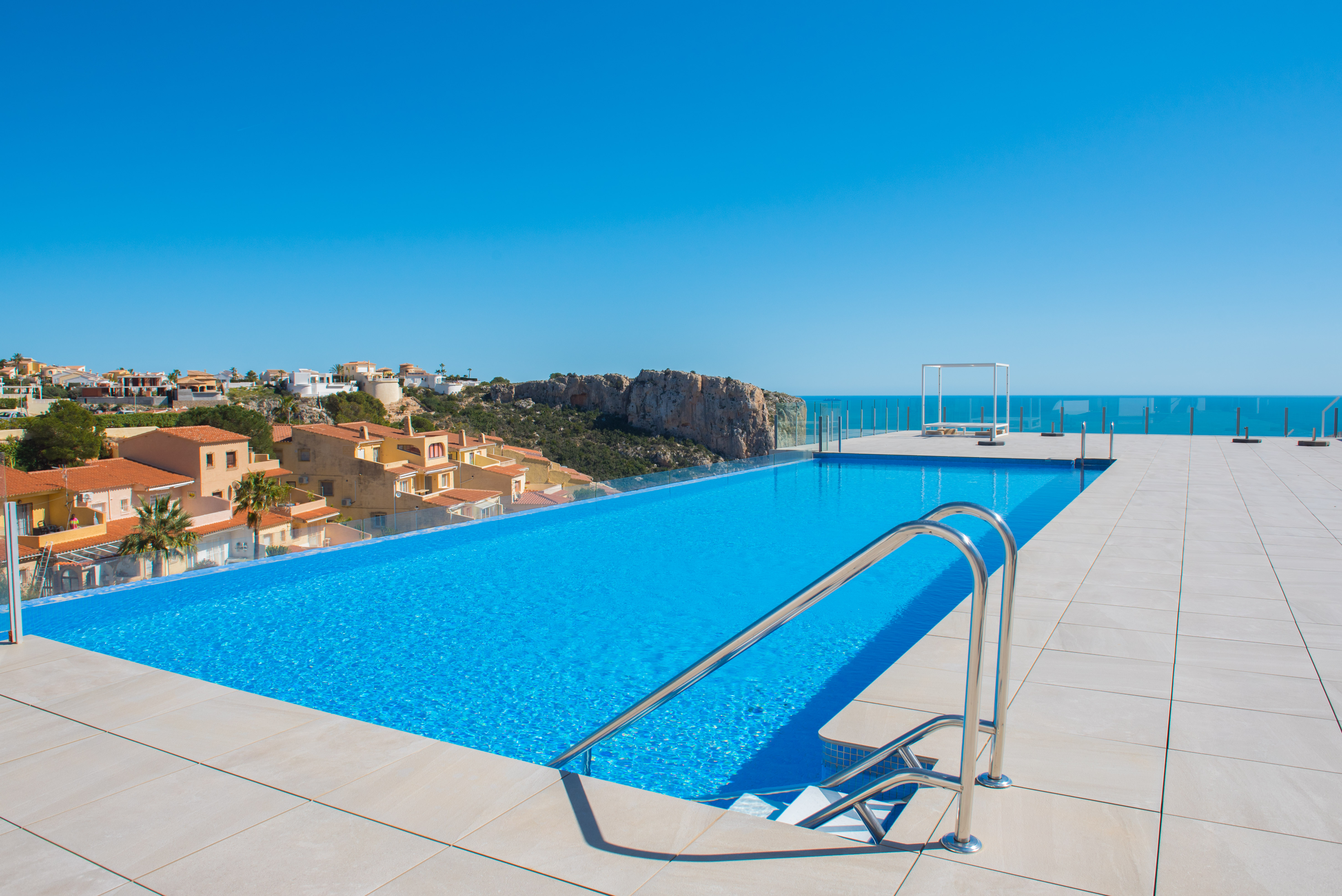 Gemeinschaftlicher Infinity-Pool mit Glasgeländer und Panoramablick auf das Meer und die Klippen. Die Terrasse ist mit hellen großformatigen Fliesen und einem Daybed ausgestattet.