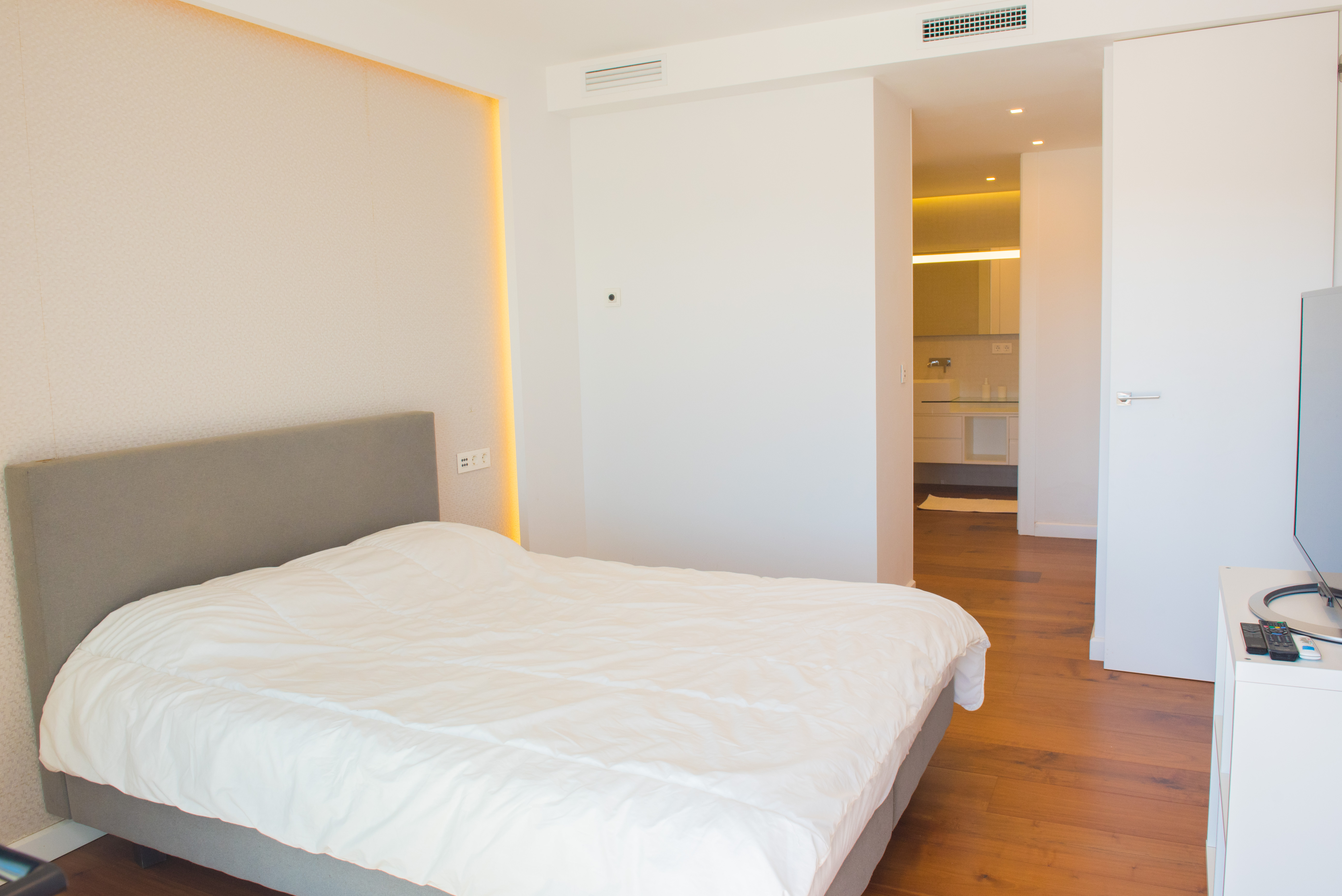 Hauptschlafzimmer mit grau gepolstertem Kopfteil, indirekter LED-Wandbeleuchtung, warmem Echtholzboden und Blick in das angrenzende, moderne Badezimmer.