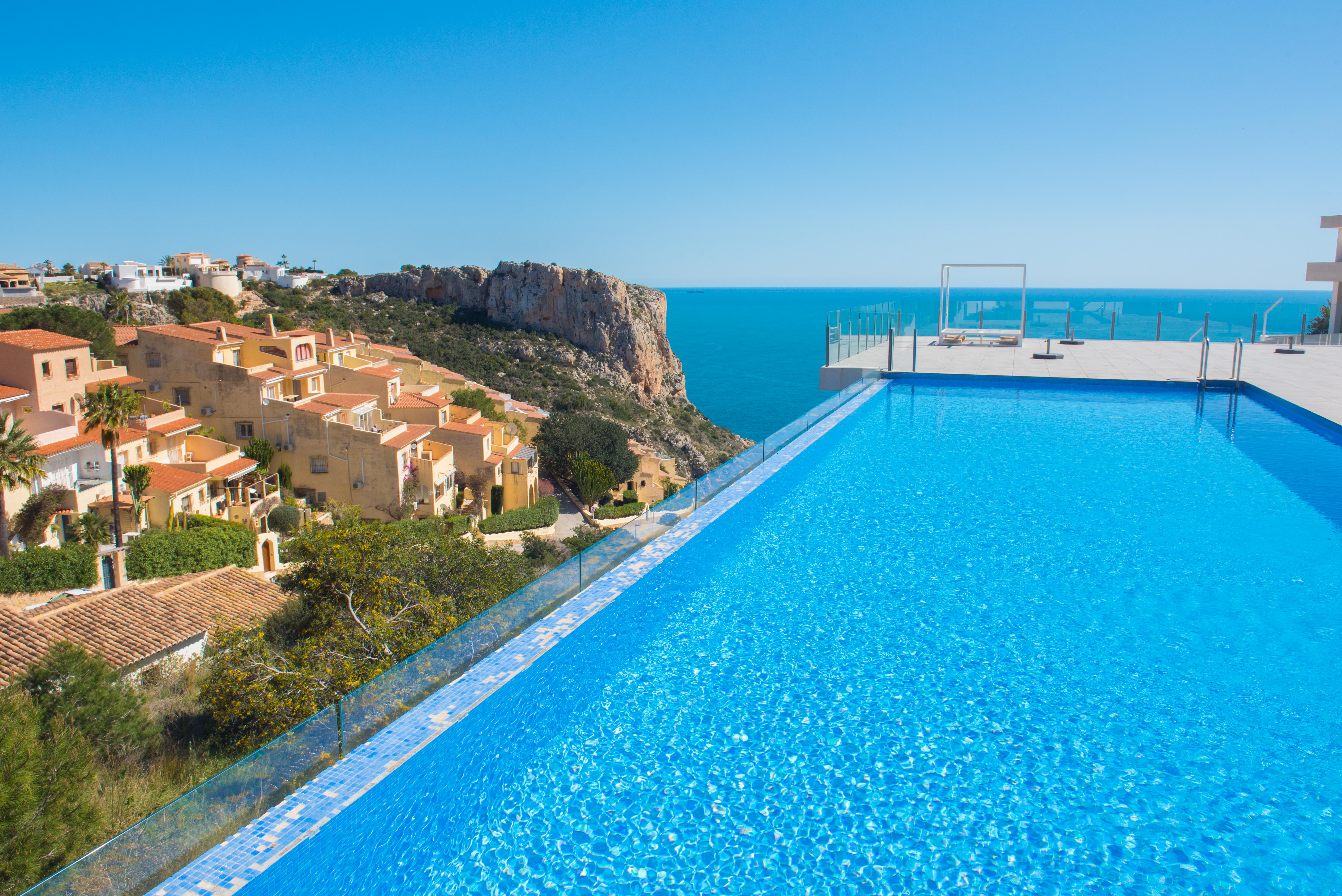 Infinity-Pool mit blauen Mosaikfliesen und Glasgeländer, mit weitem Blick über das Mittelmeer, die Klippen und die umliegende Wohnarchitektur.
