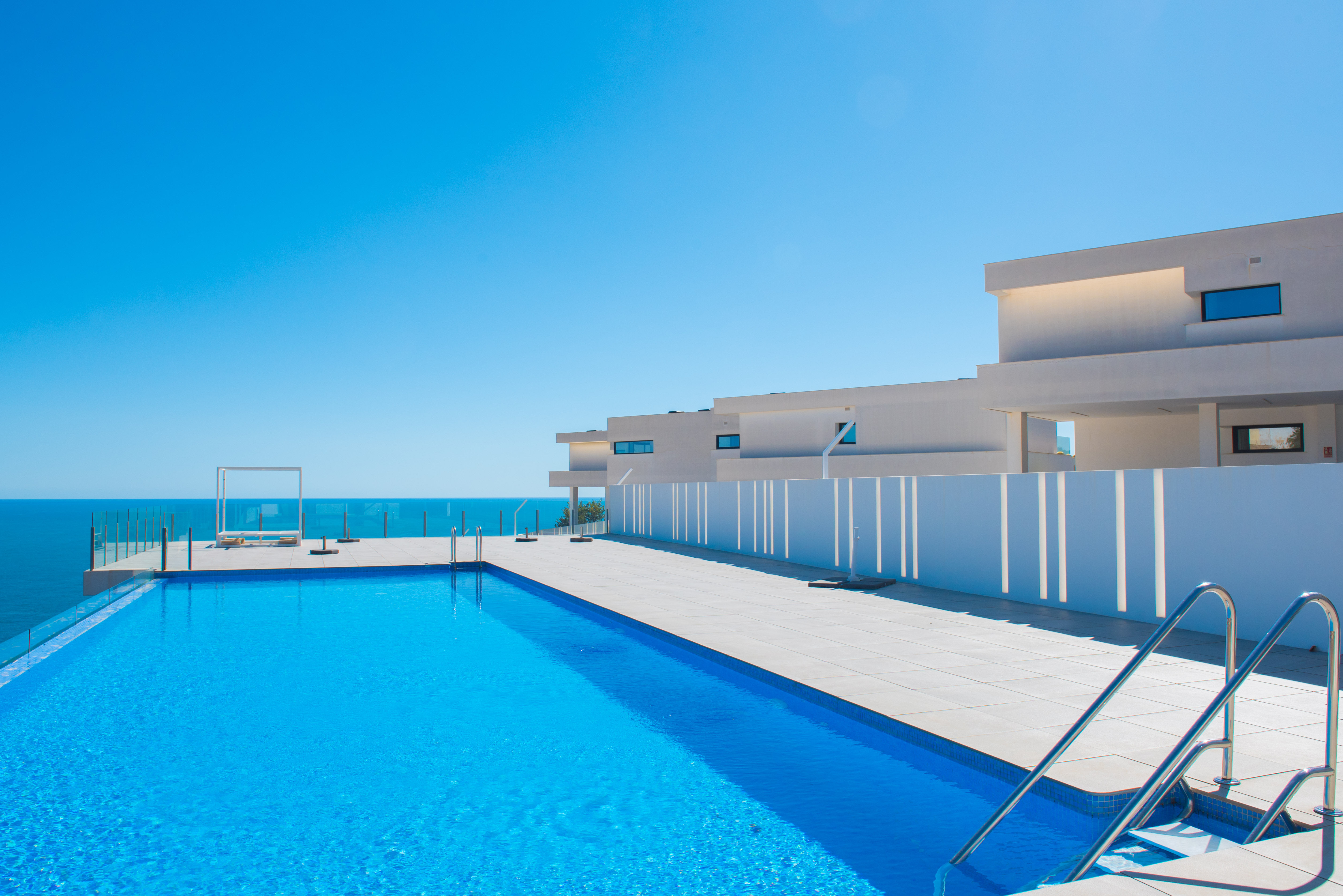 Infinity-Pool mit Blick auf das Mittelmeer, großzügiger Fliesenterrasse, Glasgeländern und minimalistischer weißer Architektur bei klarem Himmel.