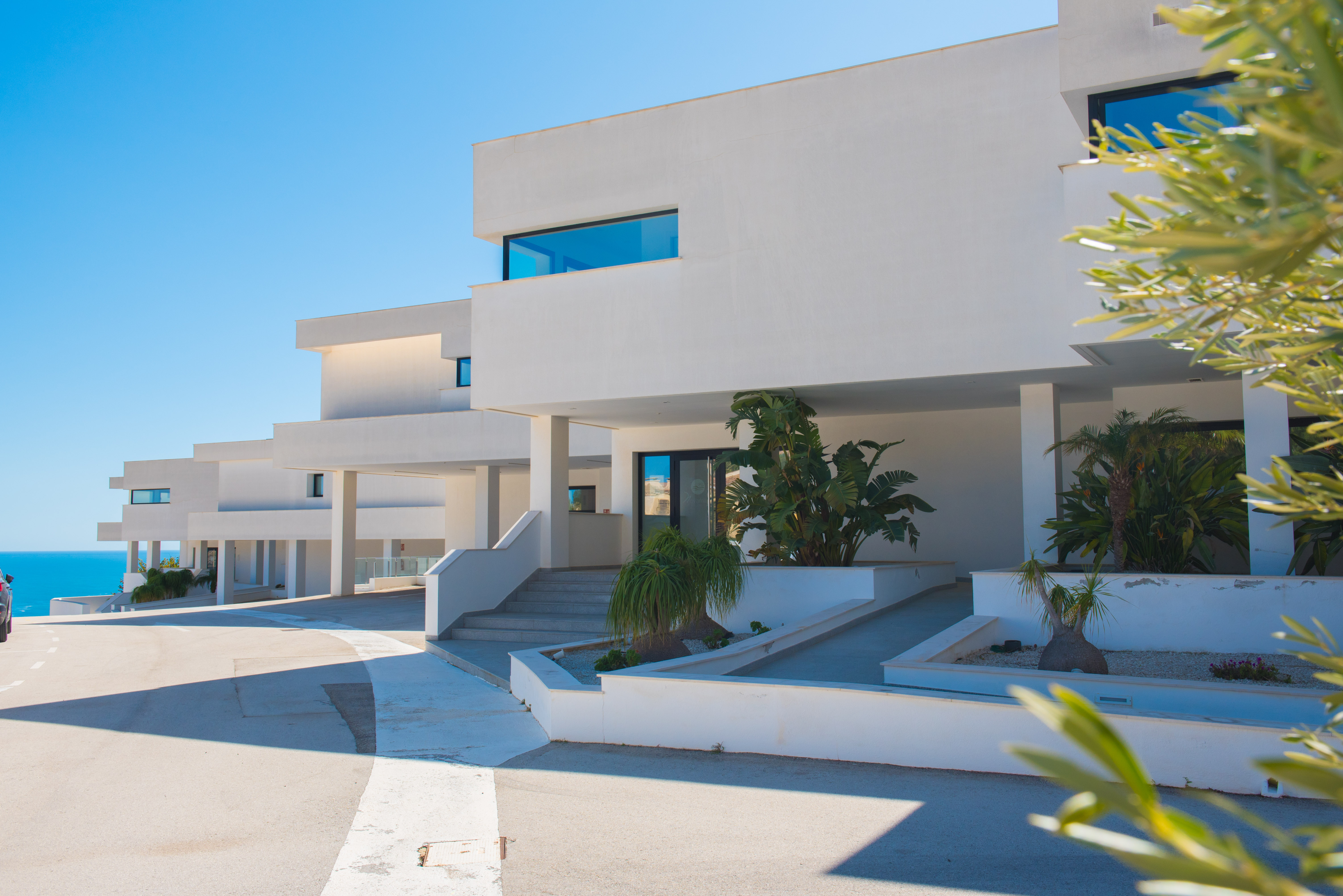 Minimalistische reinweiße Fassade mit gestuften Eingängen und Rampen, integrierten Pflanzgefäßen mit tropischem Grün und klaren Linien.