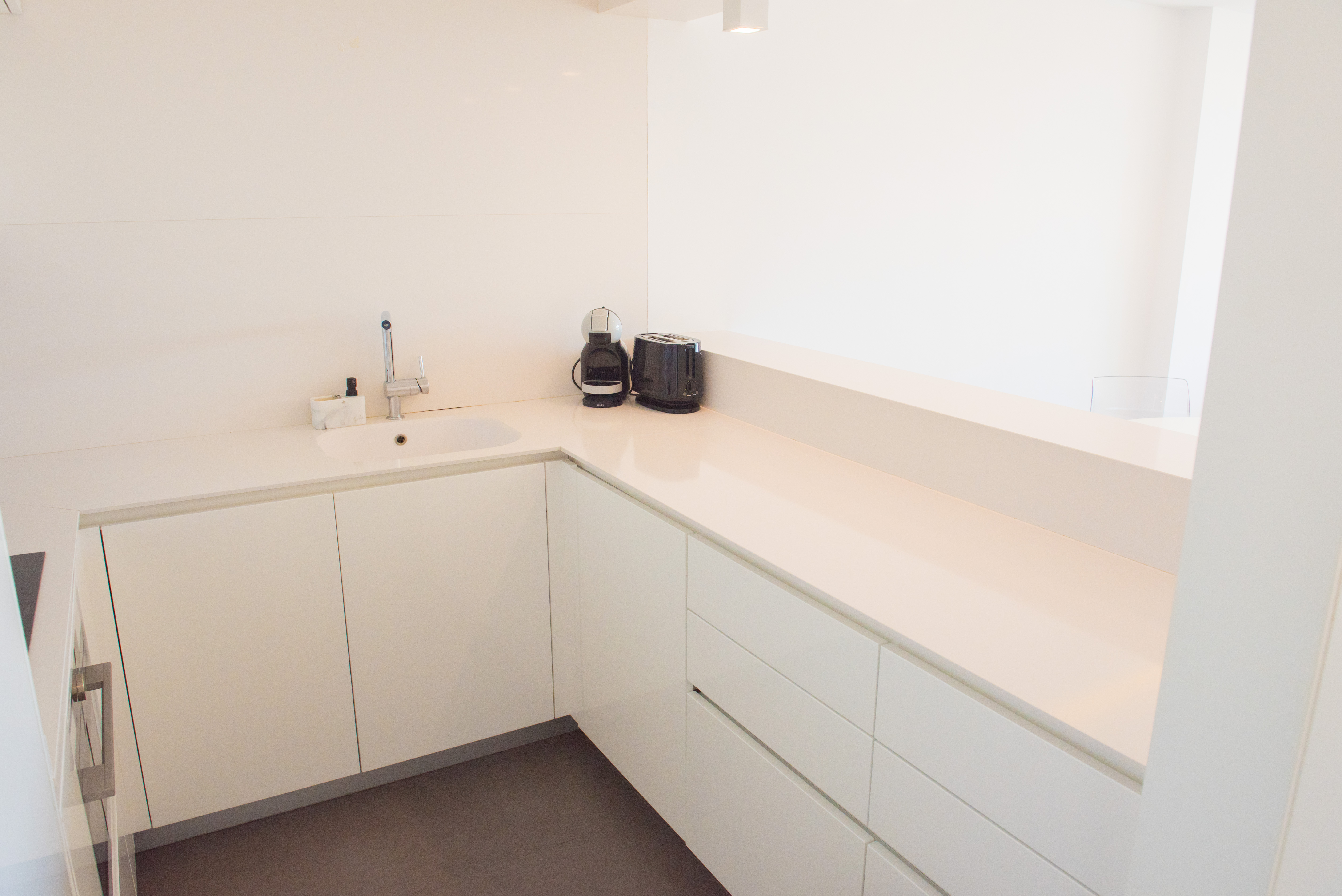 Moderne minimalistische Küche in Hochglanzweiß mit Quarz-Arbeitsplatte, integrierter Spüle und dunklem Fliesenboden für klare Linien.