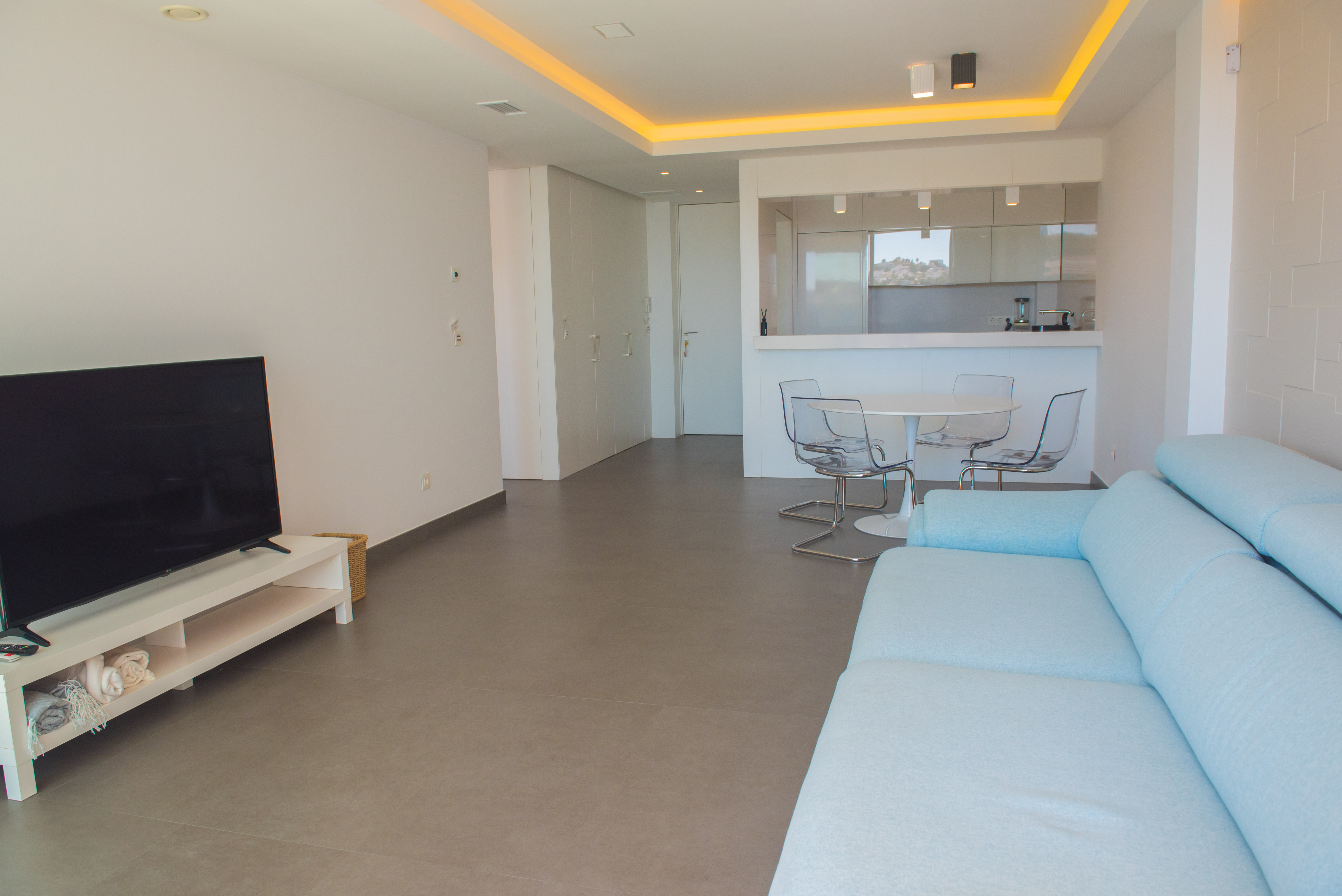 Offener Wohnbereich mit hellblauem Sofa, Essplatz mit transparenten Stühlen und integrierter Küche mit indirekter Deckenbeleuchtung.