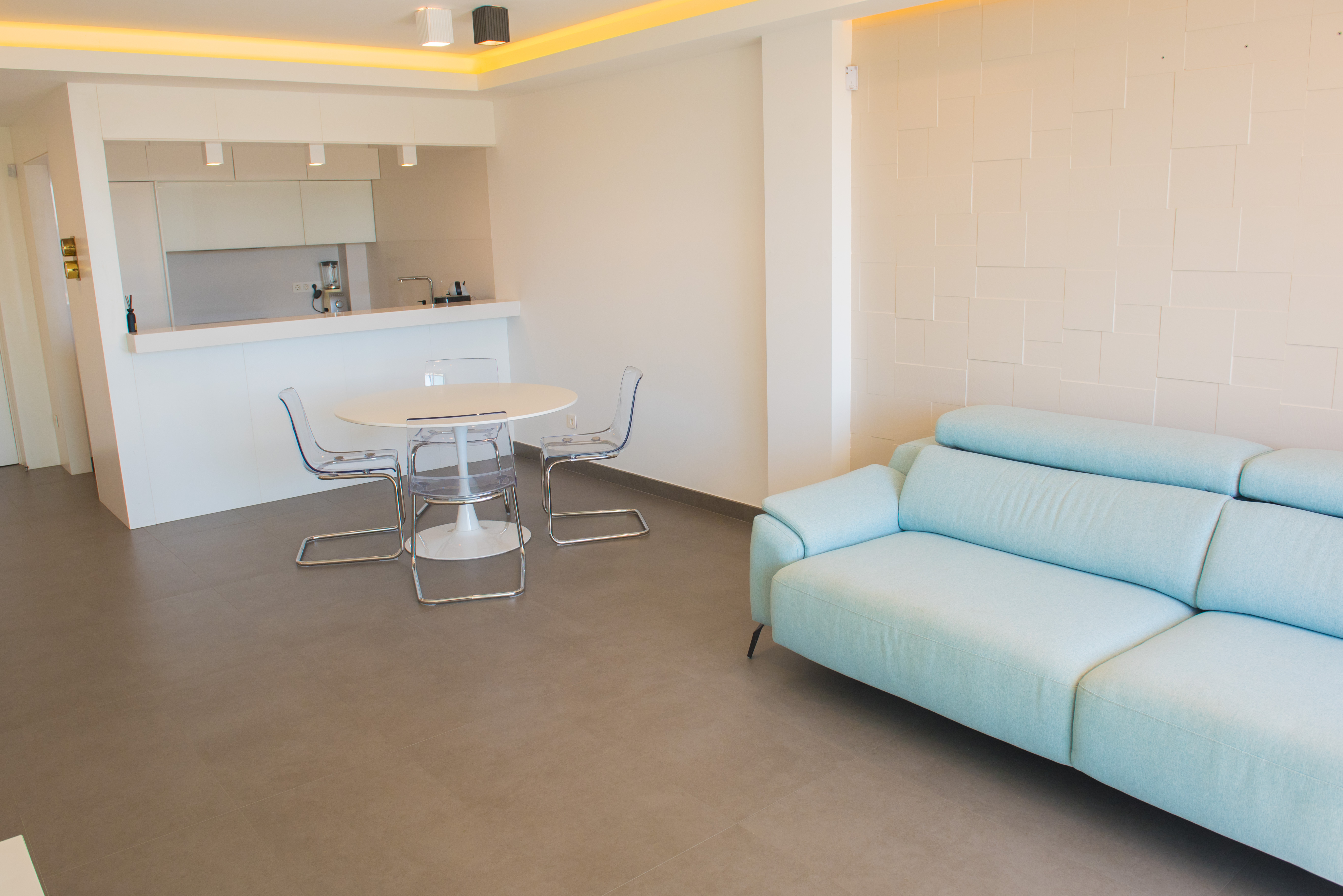 Offener Wohnbereich mit hellblauem Sofa, transparenten Acrylstühlen und weißem Esstisch. Akzentuiert durch eine geometrische Reliefwand und indirekte Deckenbeleuchtung.