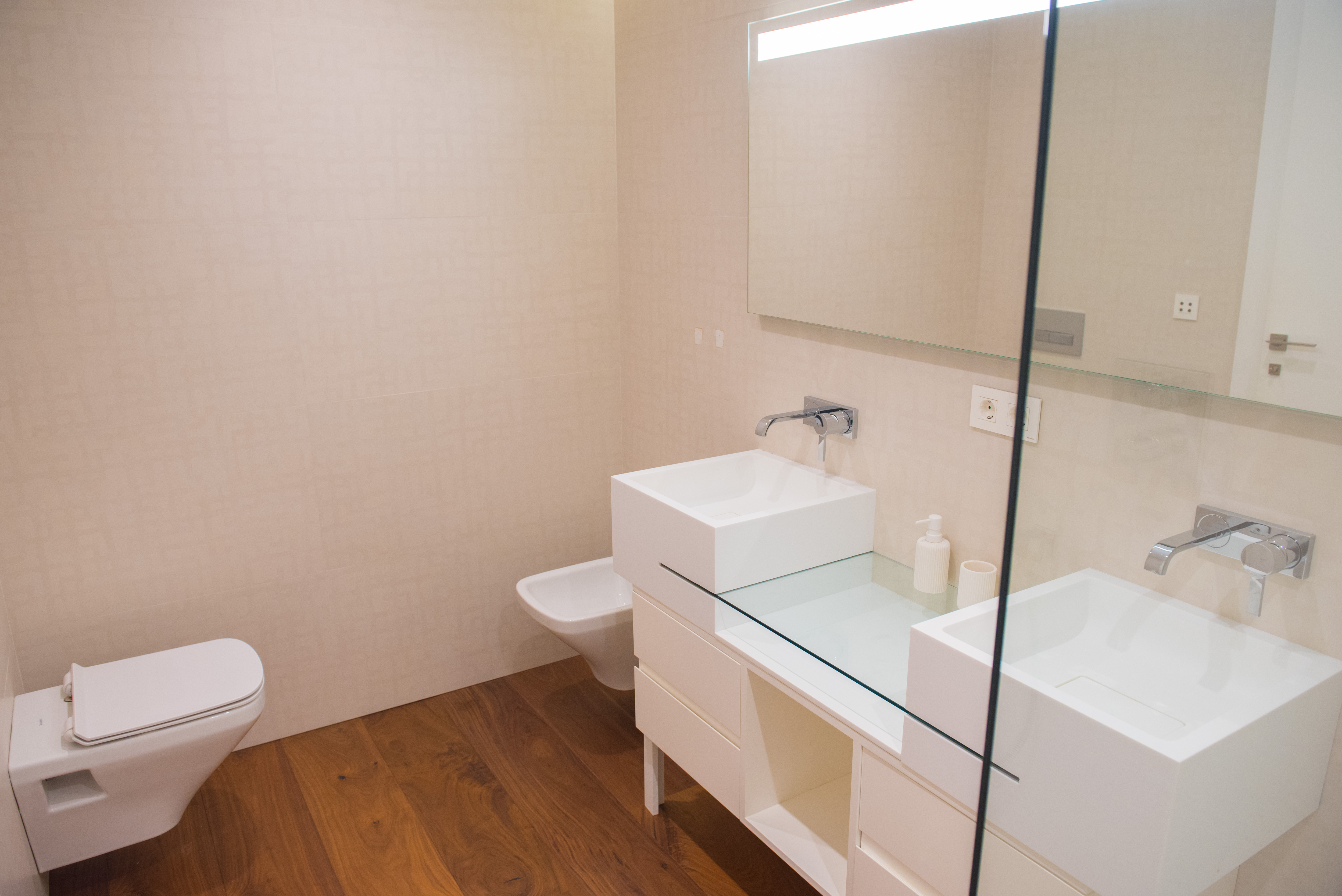 Zeitgenössisches Badezimmer mit Doppelwaschtisch, quadratischen Becken und Glasplatte. Mit wandhängenden Sanitärobjekten und hochwertigem Holzboden.