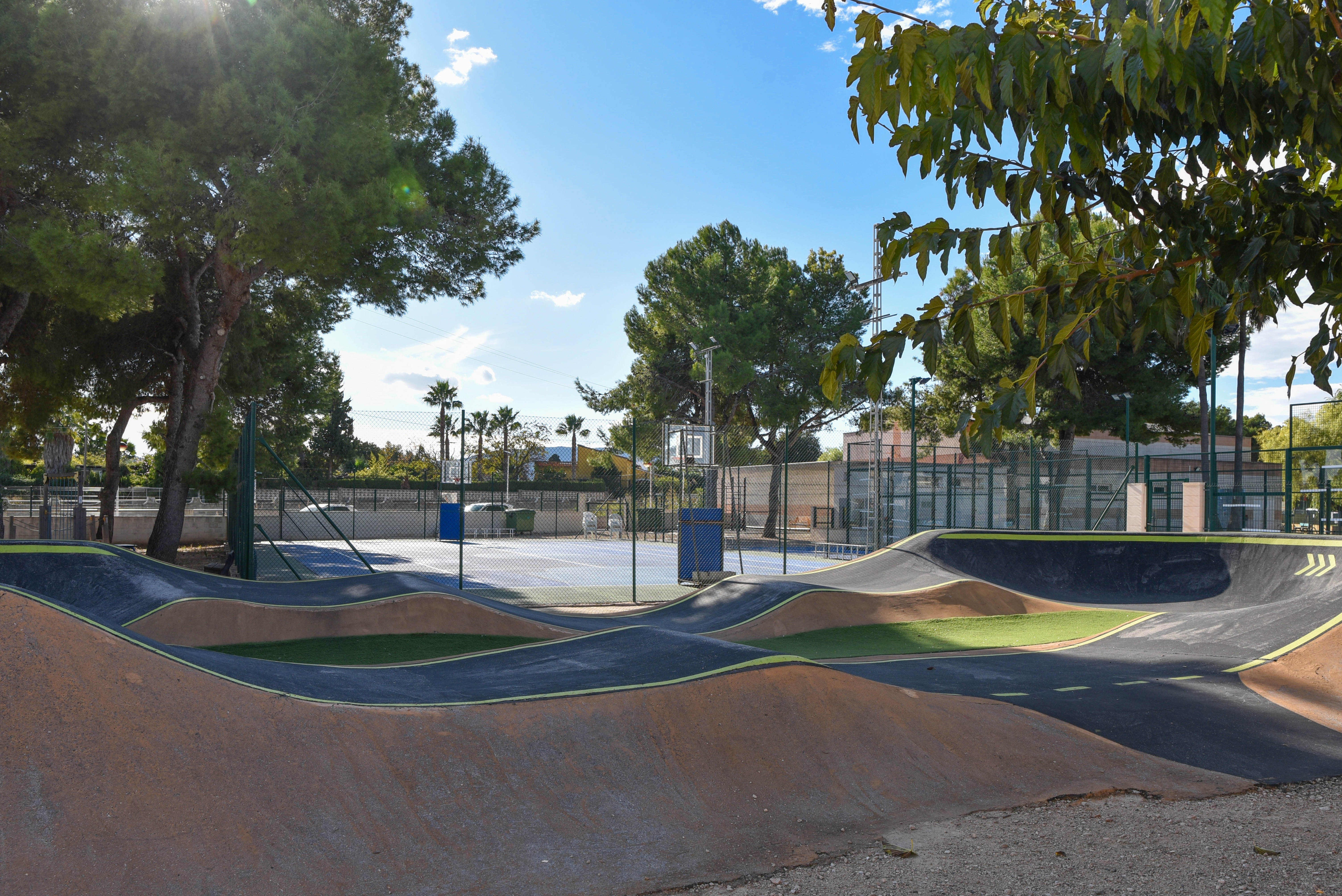 Asphaltierte Pump-Track-Anlage mit Wellenbahnen neben einem eingezäunten Multisportplatz, umgeben von Pinien in einem städtischen Erholungsgebiet.