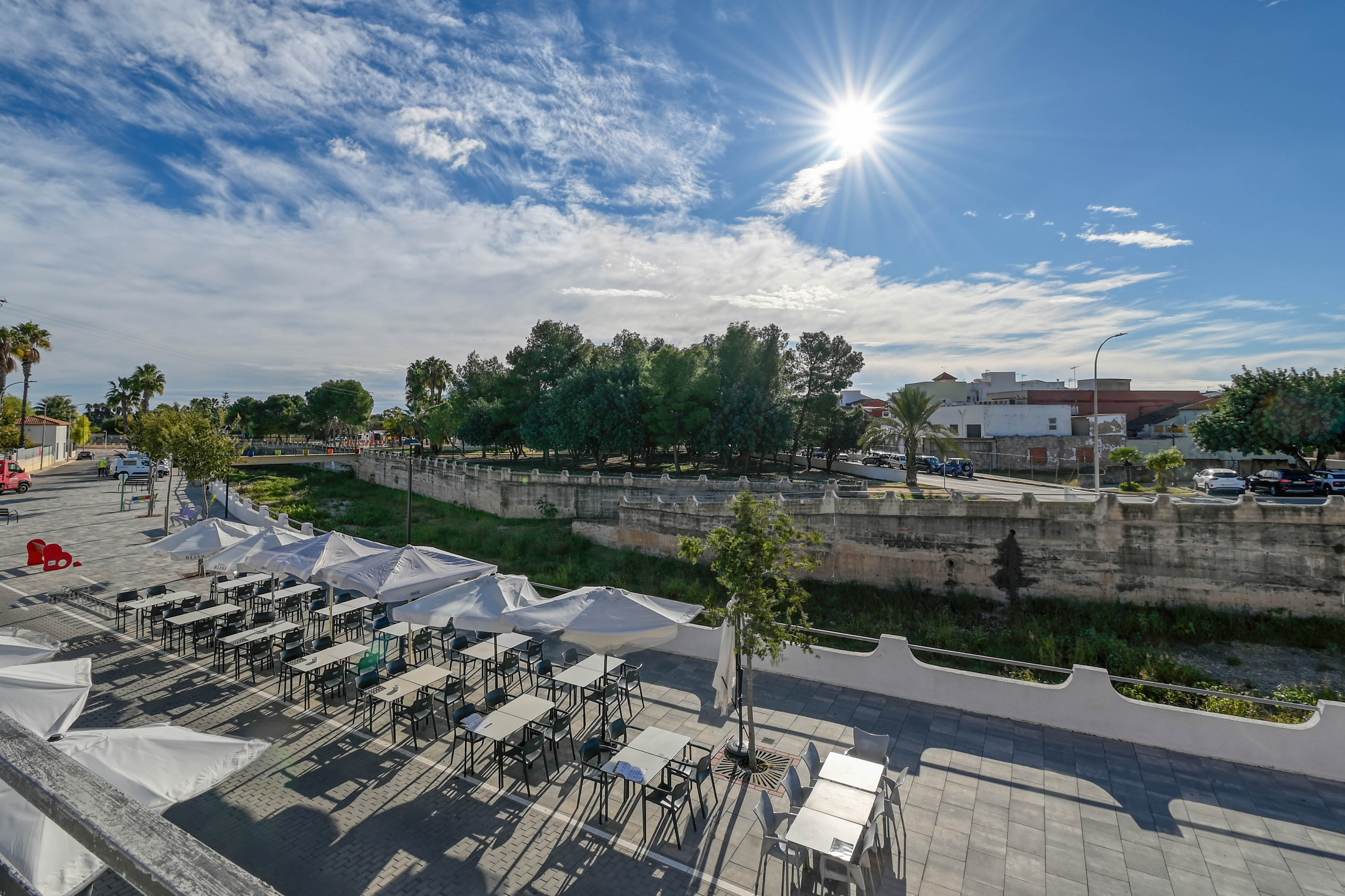 Außenbereich einer Terrasse mit weißen Sonnenschirmen und Tischen, mit Blick auf einen steinernen Kanalwall und eine parkähnliche Anlage bei Sonnenschein.