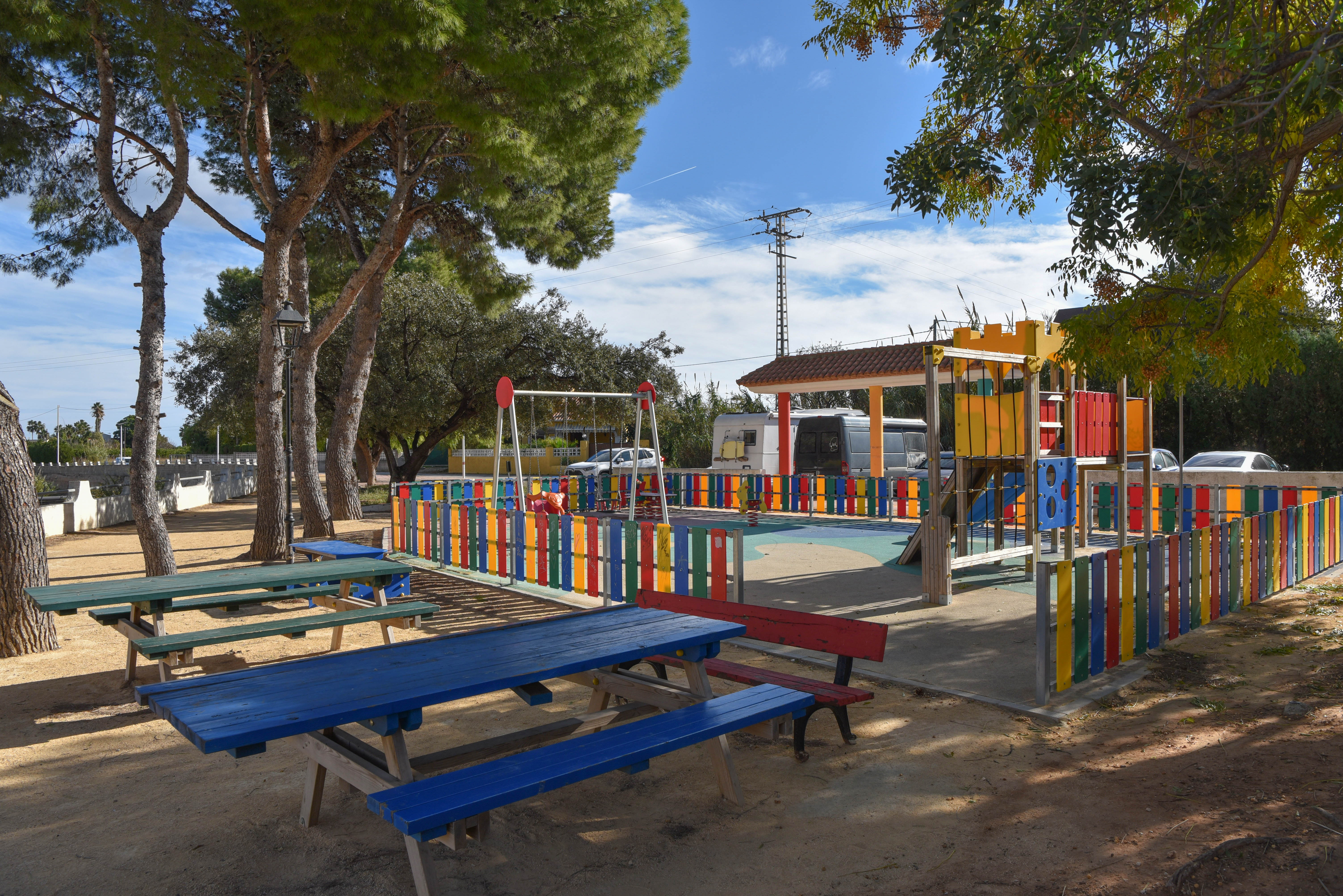 Eingezäunter Kinderspielplatz mit bunten Spielgeräten und hölzernen Picknicktischen im Schatten alter Pinien in einer gepflegten Außenanlage.