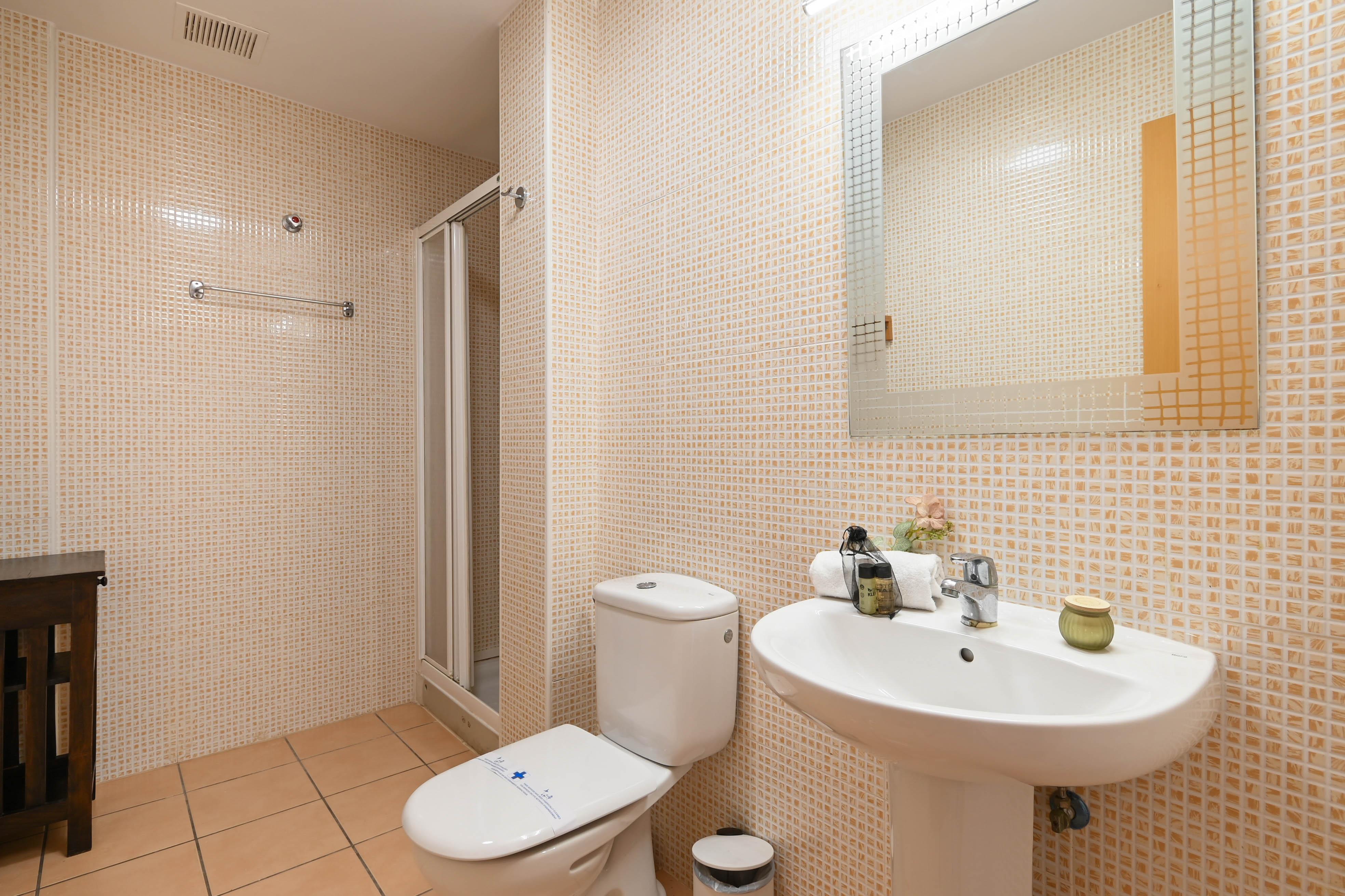 Funktionales Badezimmer mit beigen Mosaikwandfliesen, einem weißen Standwaschbecken und einer Dusche mit Glasfalttür.