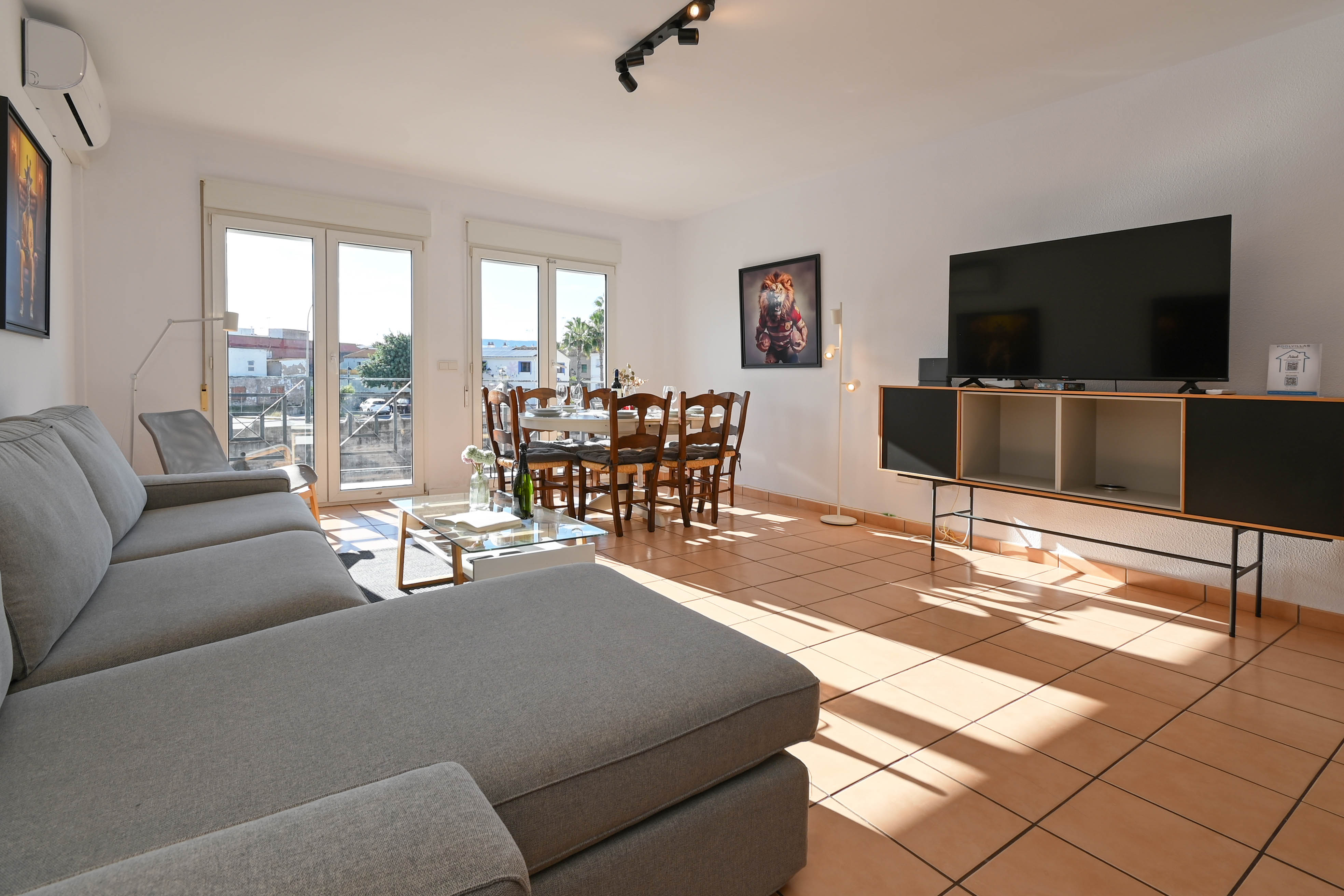 Heller, offener Wohn- und Essbereich mit Terrakottaboden, großem grauem Ecksofa und direktem Zugang zum Balkon durch bodentiefe Glastüren.