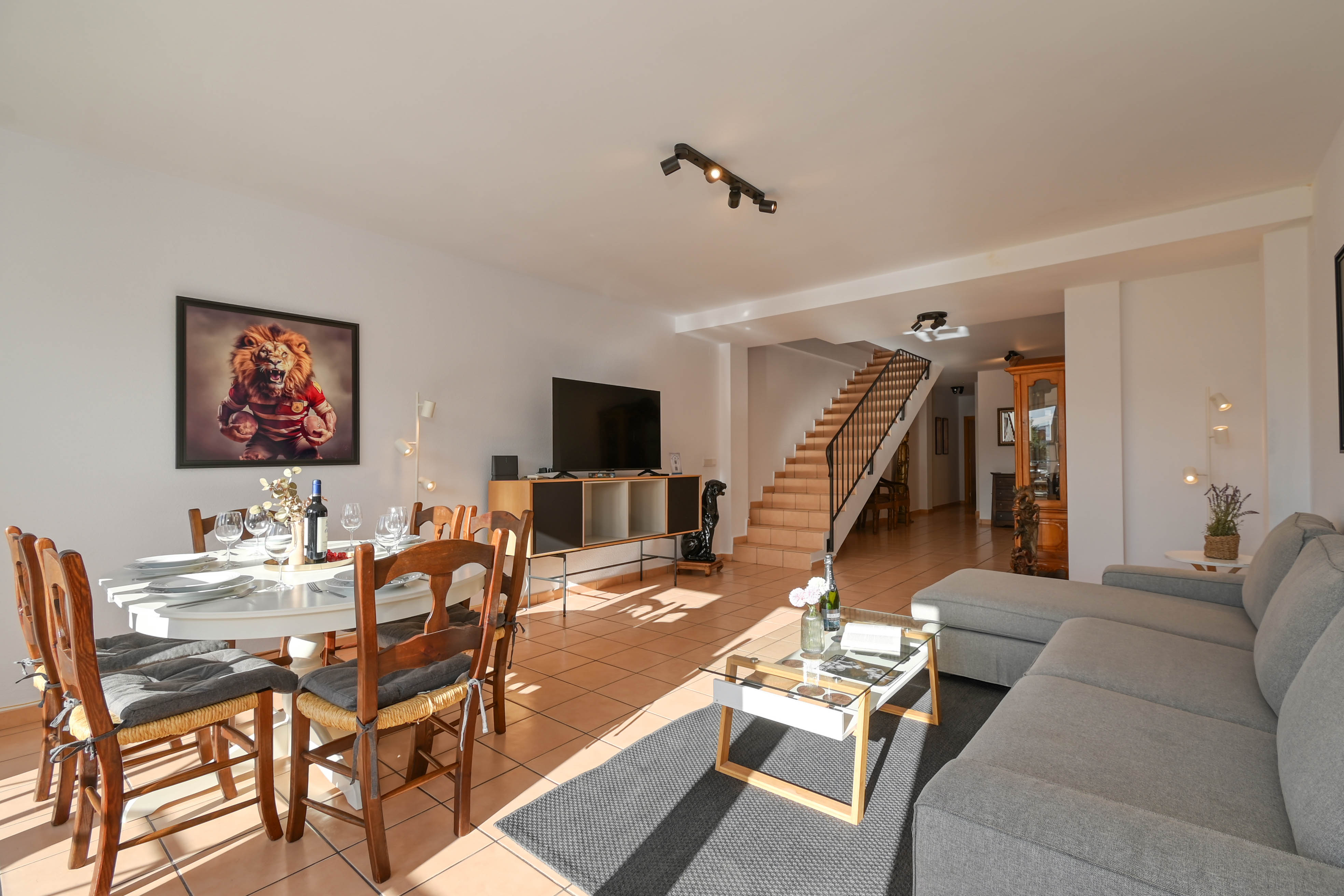Offener Wohn- und Essbereich mit Terrakottafliesen, grauem L-Sofa und einer Treppe mit Metallgeländer, die in die obere Etage führt.