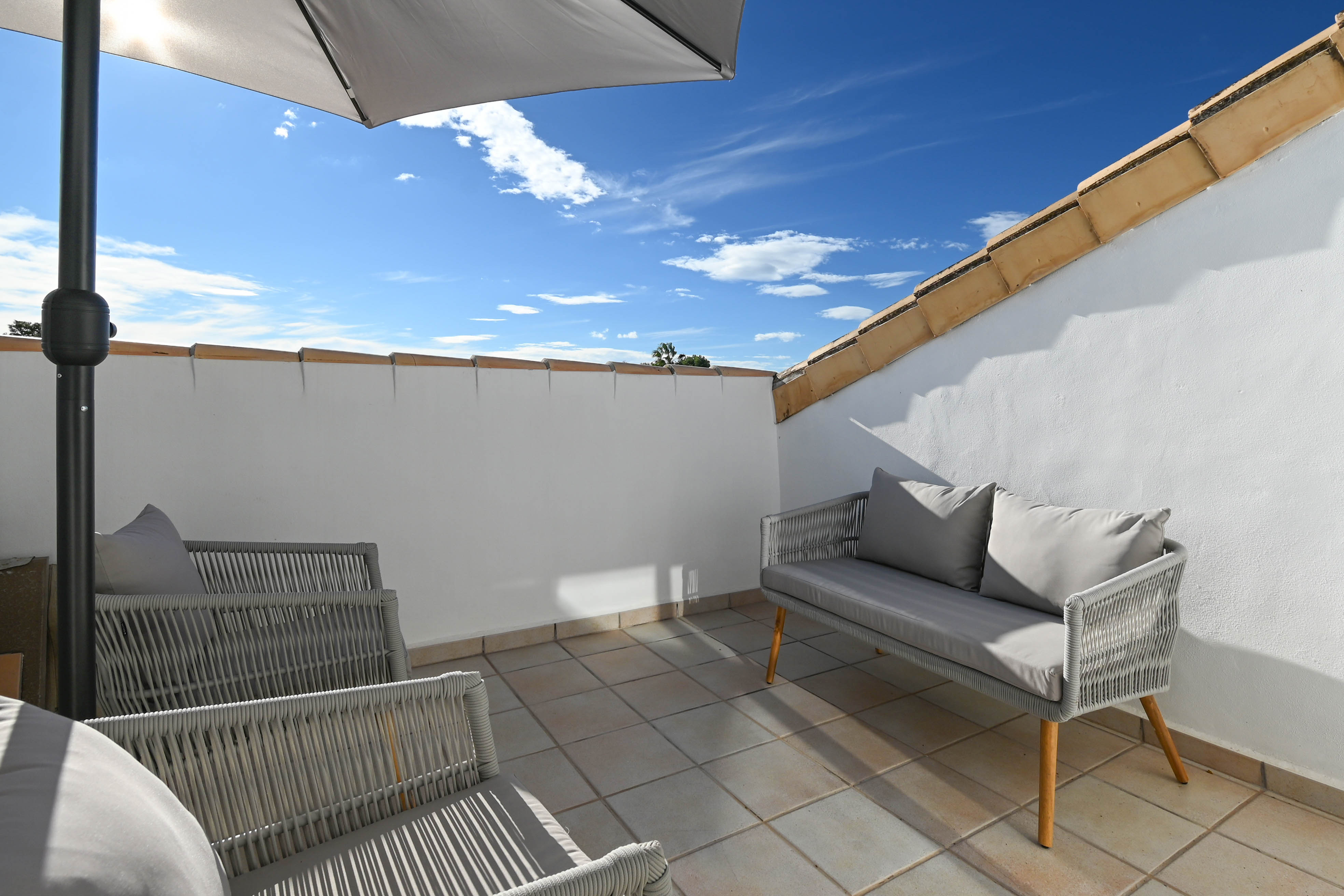 Private Dachterrasse mit grauen Outdoor-Möbeln, Fliesenboden und Sonnenschirm, umrahmt von weißen Wänden und traditionellen Dachziegeln.