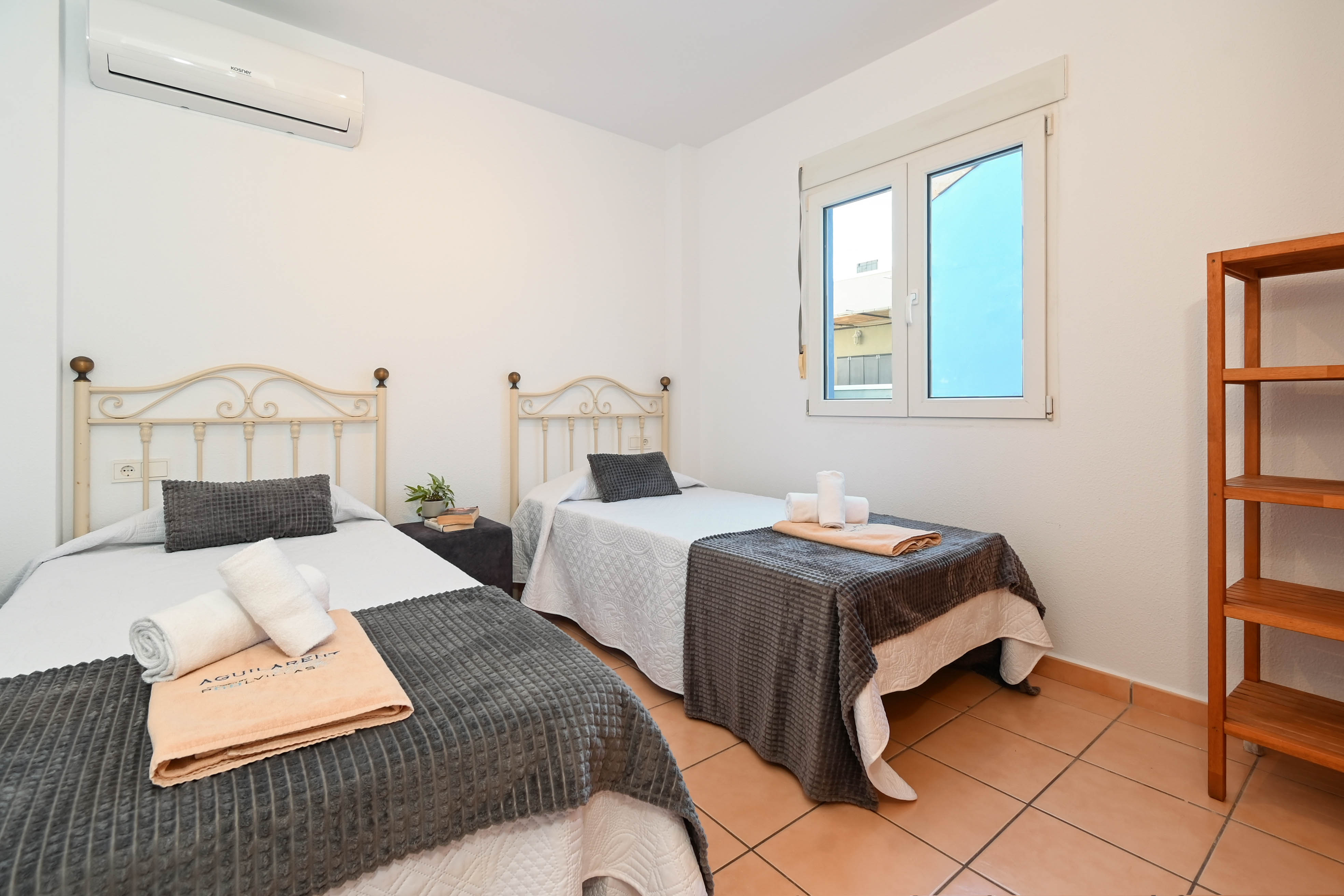 Schlafzimmer mit zwei Einzelbetten, schmiedeeisernen Kopfteilen, Terrakotta-Fliesenboden und Klimaanlage. Mit Holzregal und weiß gerahmtem Fenster für Tageslicht.