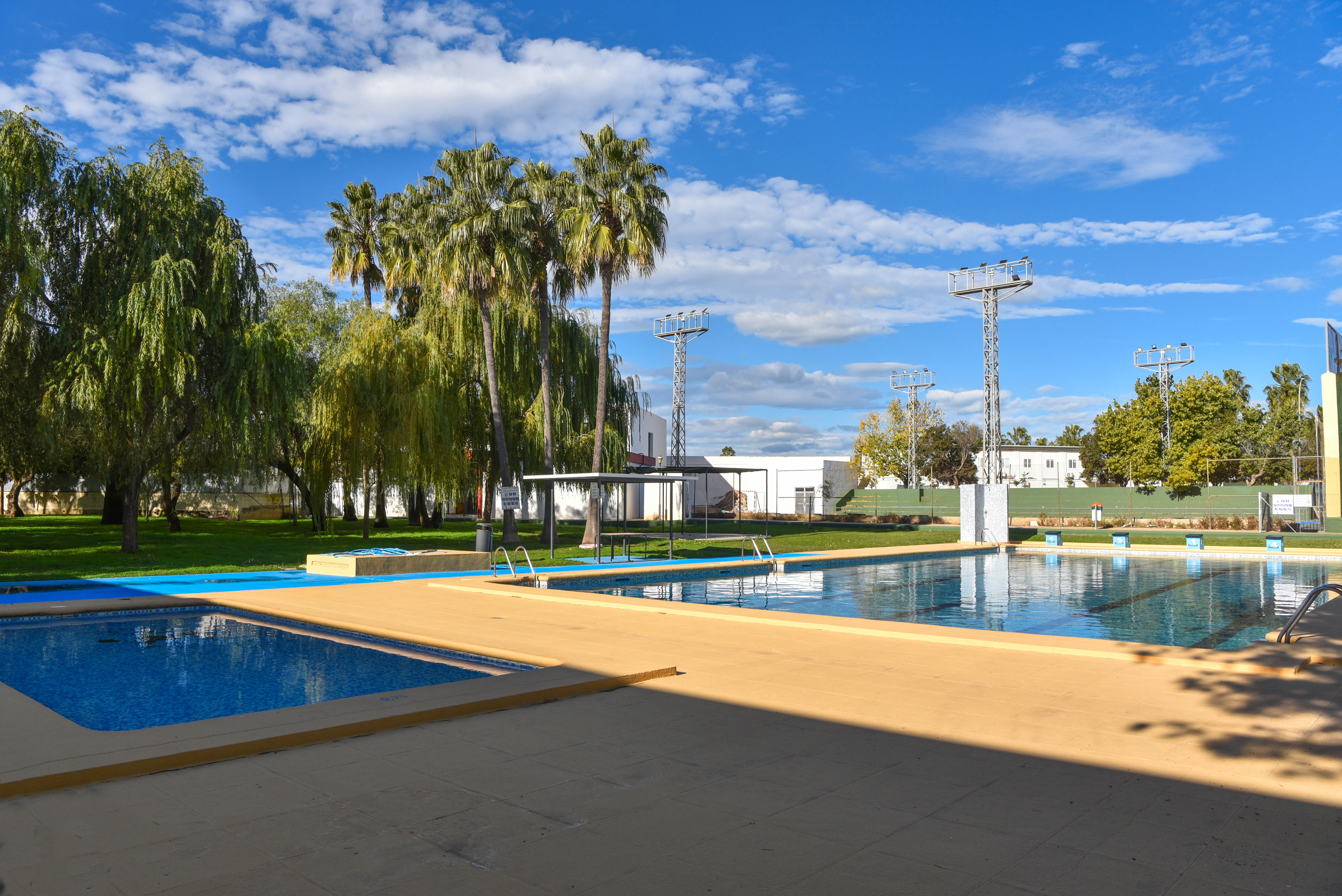 Städtisches Schwimmbad mit Hauptbecken, Kinderbecken, Liegeterrasse und Grünanlage mit Palmen sowie Flutlichtmasten für den Sportbereich.