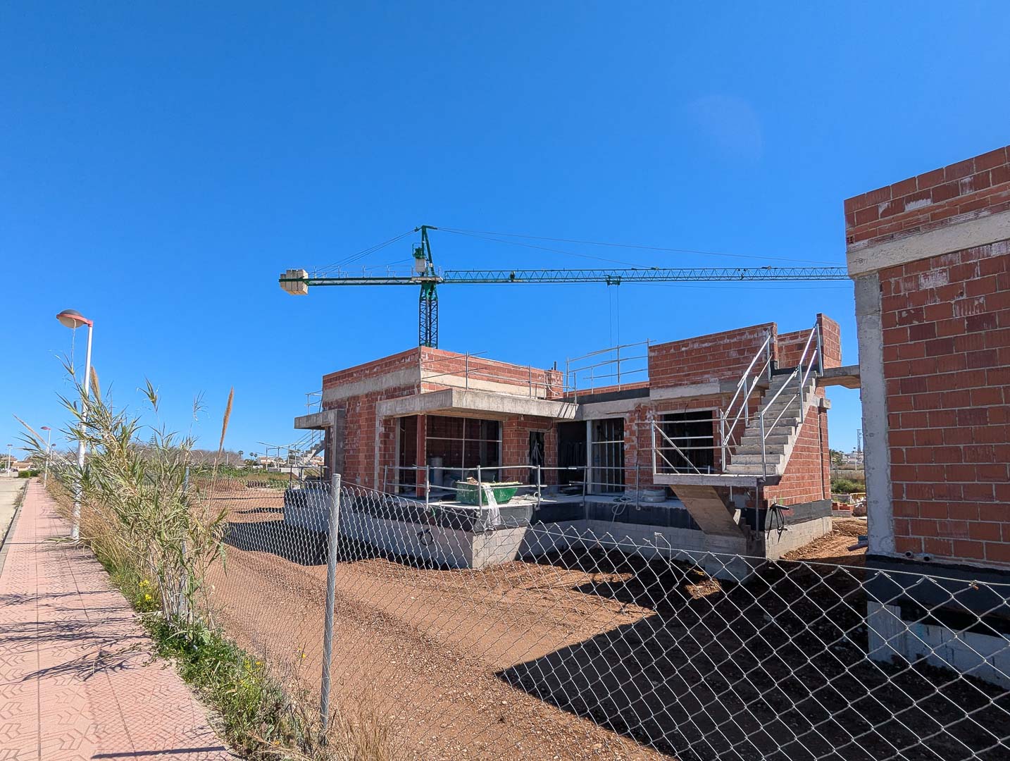 Aktueller Baustatus mit rotem Ziegelmauerwerk, Betonplatten und Außentreppe auf einem flachen Grundstück unter blauem Himmel mit Baukran.