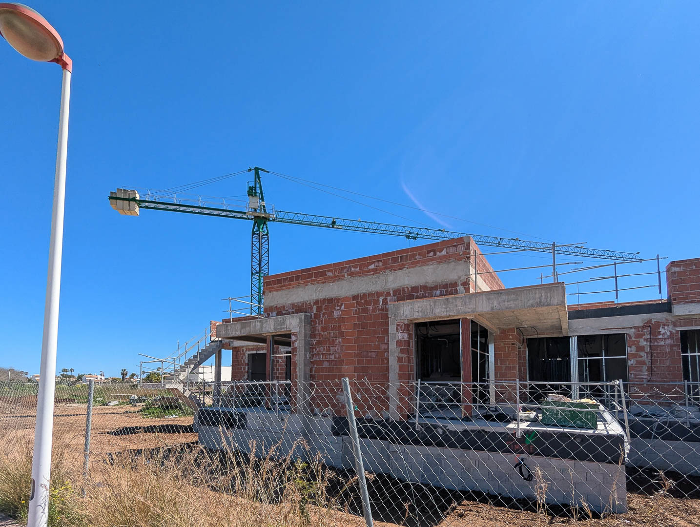 Aktueller Stand der Bauarbeiten mit Ziegel- und Betonrohbau, inklusive Baukran und Umzäunung des Grundstücks unter strahlend blauem Himmel.