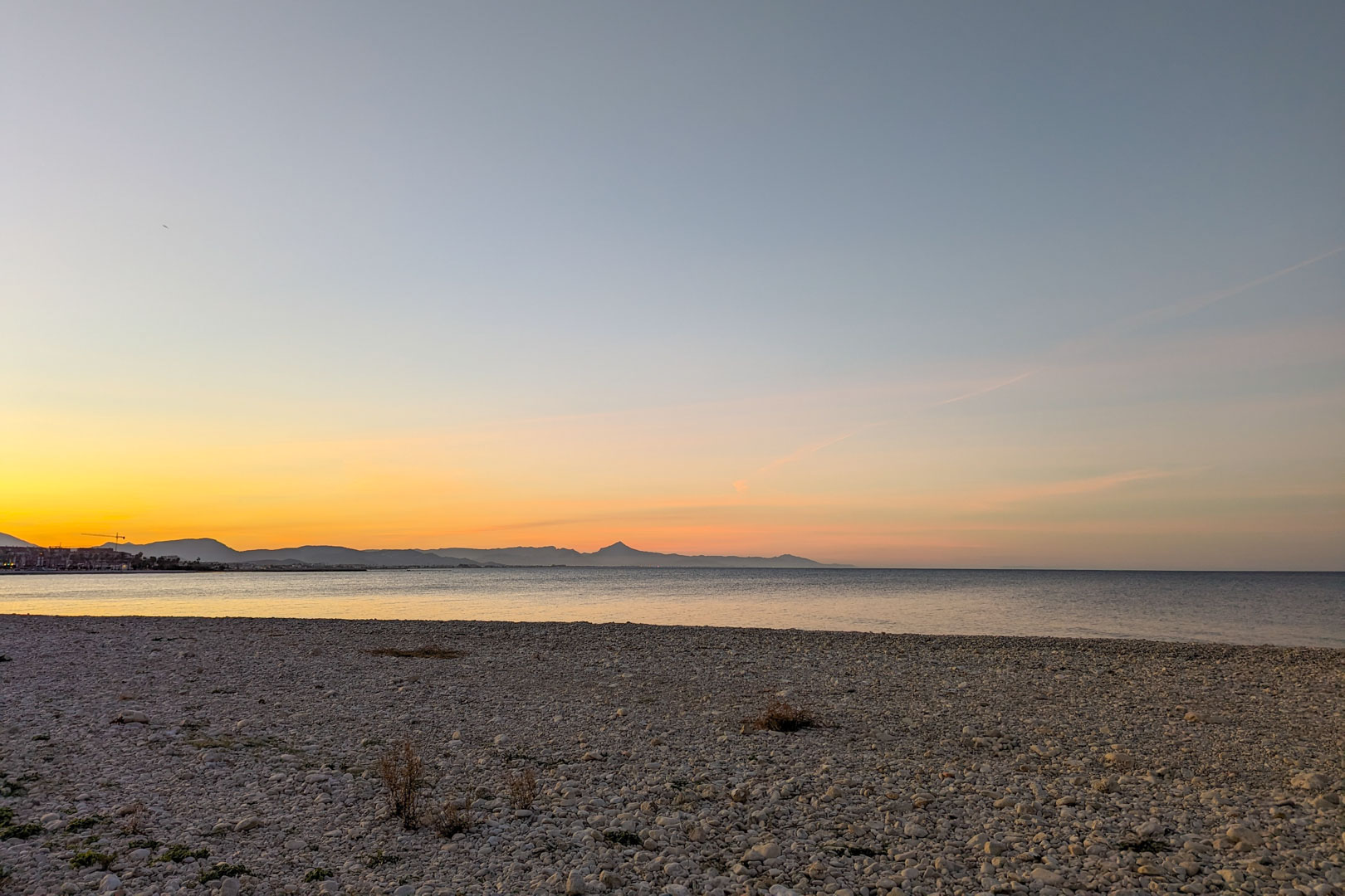 Panoramablick auf den nahegelegenen Kieselstrand bei Sonnenuntergang, mit Bergsilhouetten am Horizont und ruhigem Mittelmeerwasser.