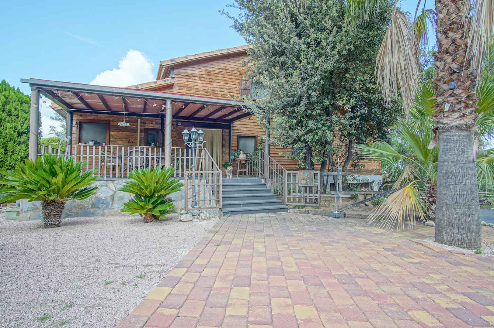 Außenansicht aus finnischem Kiefernholz mit erhöhter Veranda, Natursteinsockel und gepflastertem Zugangsweg inmitten mediterraner Bepflanzung.