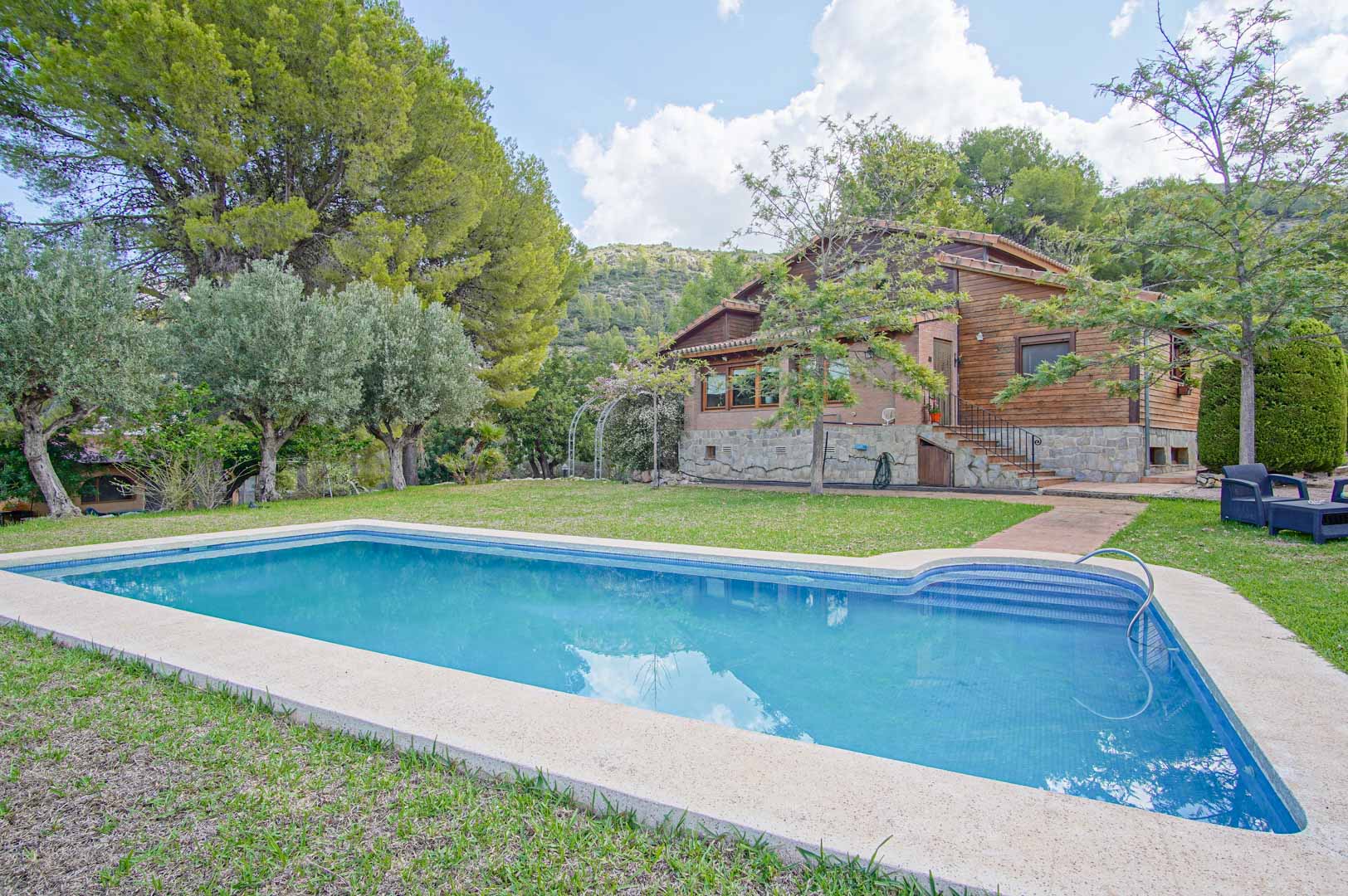 Außenansicht der Villa mit rechteckigem Pool, umgeben von Rasen. Die Fassade aus finnischem Kiefernholz und Naturstein fügt sich harmonisch in die bergige Landschaft ein.