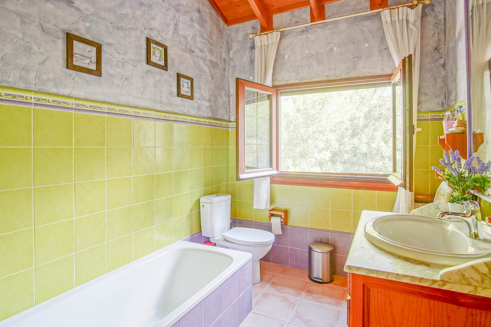 Badezimmer mit sichtbaren Holzbalken, grünen Wandfliesen und einem Waschtisch mit Marmorplatte. Ein großes Fenster bietet reichlich Tageslicht und Ausblick in die Natur.