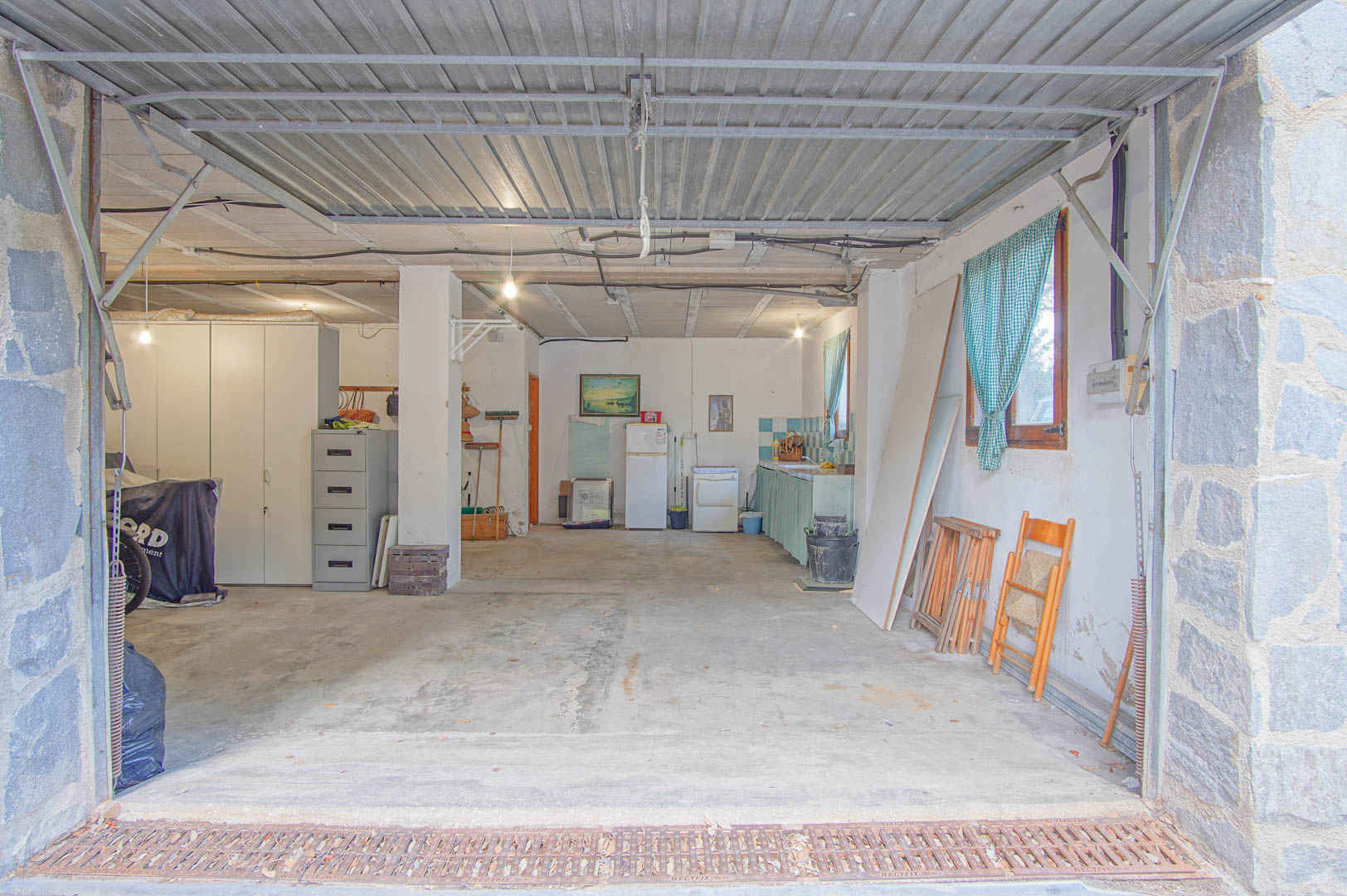 Geräumiges Souterrain mit Platz für mehrere Fahrzeuge, Werkstattbereich und integrierter Küchenzeile. Es verfügt über Betonböden, Natursteinpfeiler und ein Metall-Schwingtor.