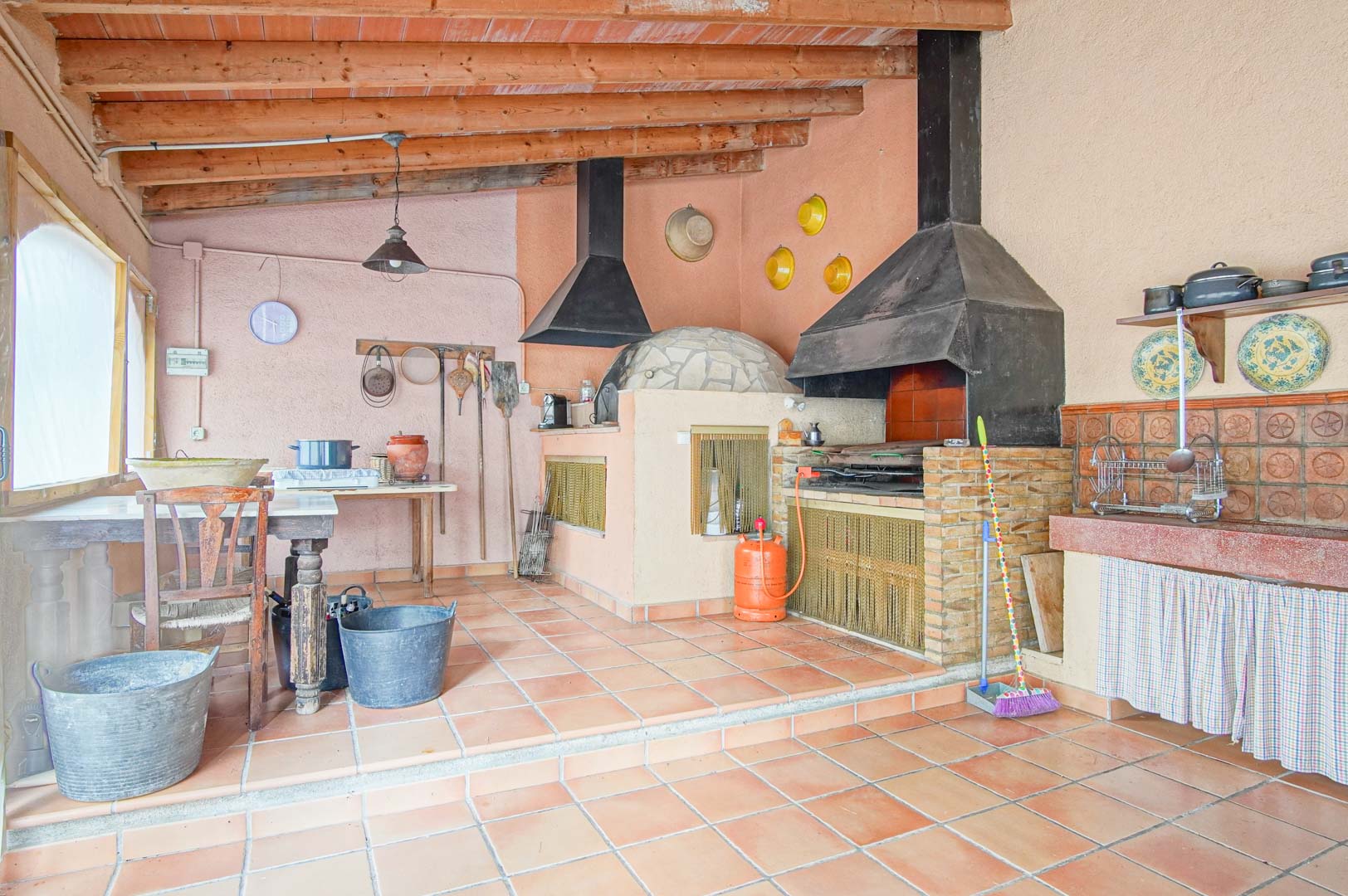 Rustikale Außenküche mit traditionellem Steinbackofen, gemauertem Grill und Sichtbalkendecke über einem klassischen Terrakottaboden.