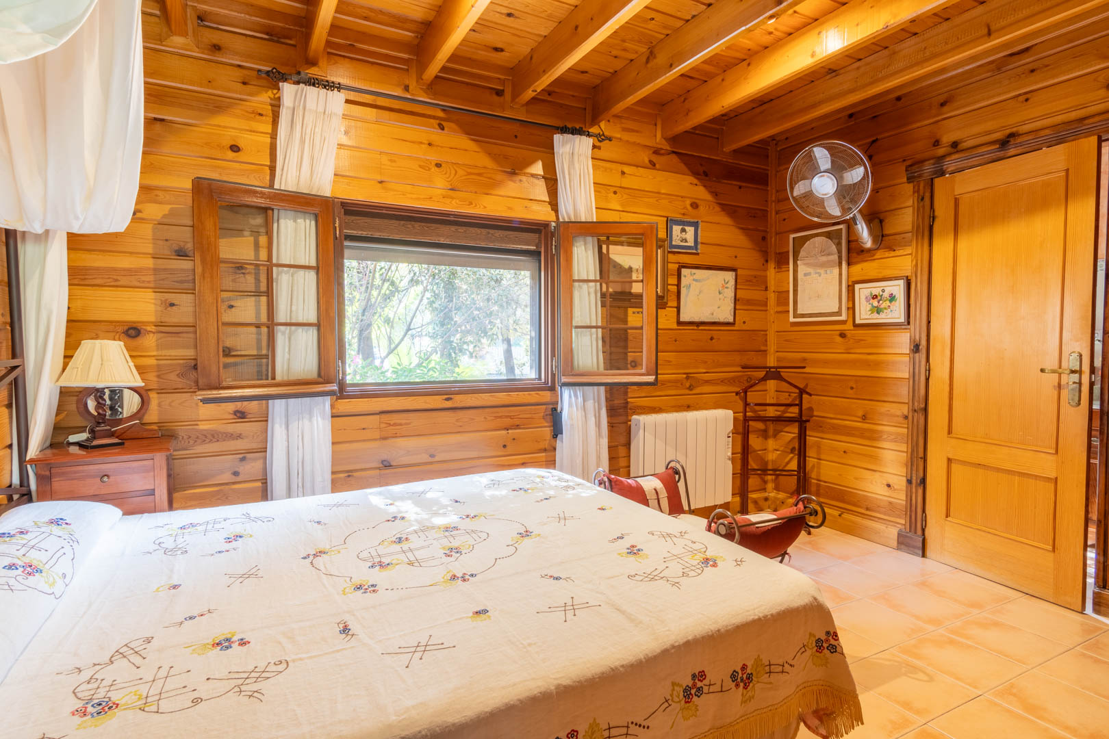 Rustikales Schlafzimmer mit finnischer Kiefernholzverkleidung, sichtbaren Deckenbalken, Fliesenboden und Holzfenstern mit Fensterläden für natürliches Licht.