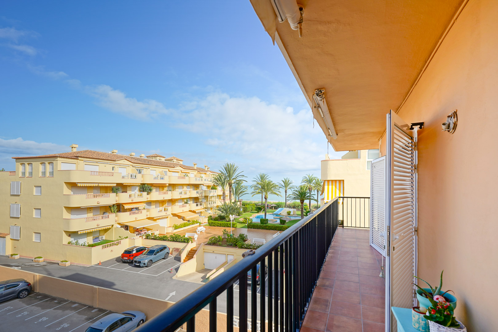 Balkon mit Terrakottafliesen und schwarzem Geländer mit Blick auf den Gemeinschaftspool, die Gartenanlagen und das Mittelmeer.
