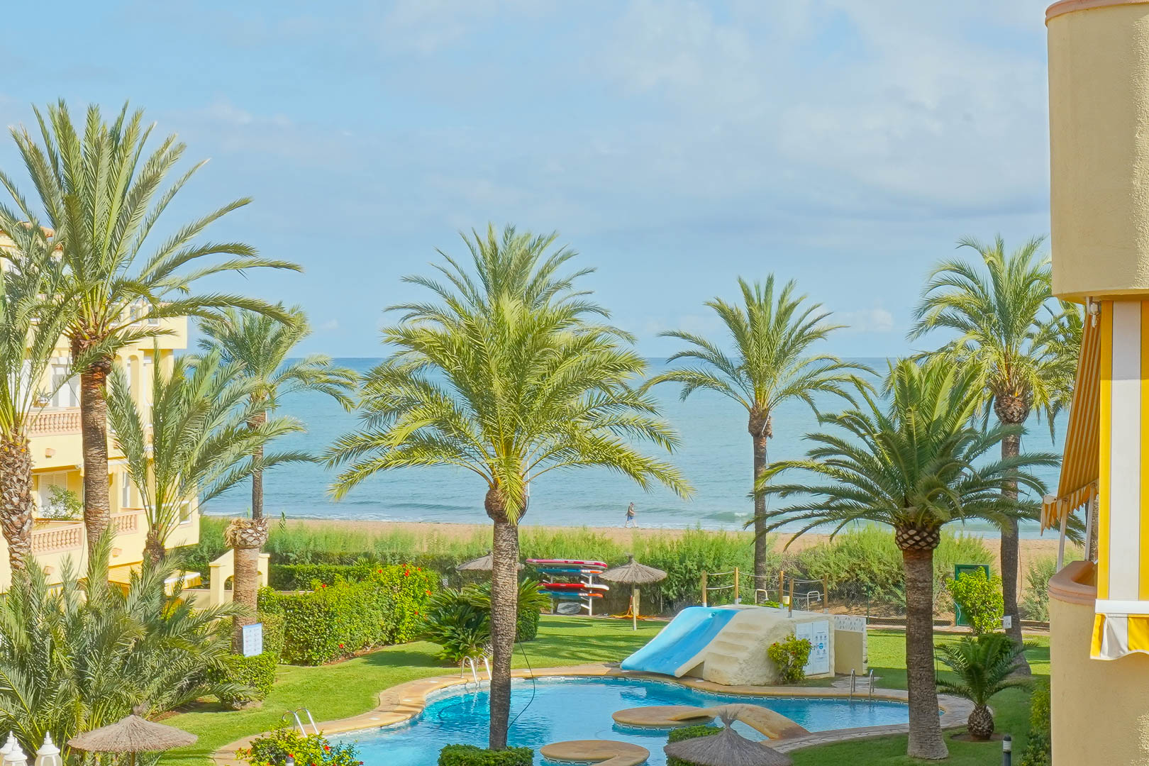 Blick auf den gemeinschaftlichen Poolbereich mit Rutsche, umgeben von Palmen und Grünflächen, mit direktem Ausblick auf den Sandstrand und das Meer.