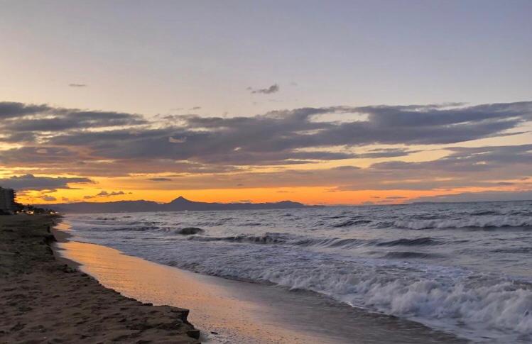 Blick auf den Sonnenuntergang über einem Sandstrand mit Brandung und Bergsilhouetten unter einem bewölkten, orangefarbenen Himmel.
