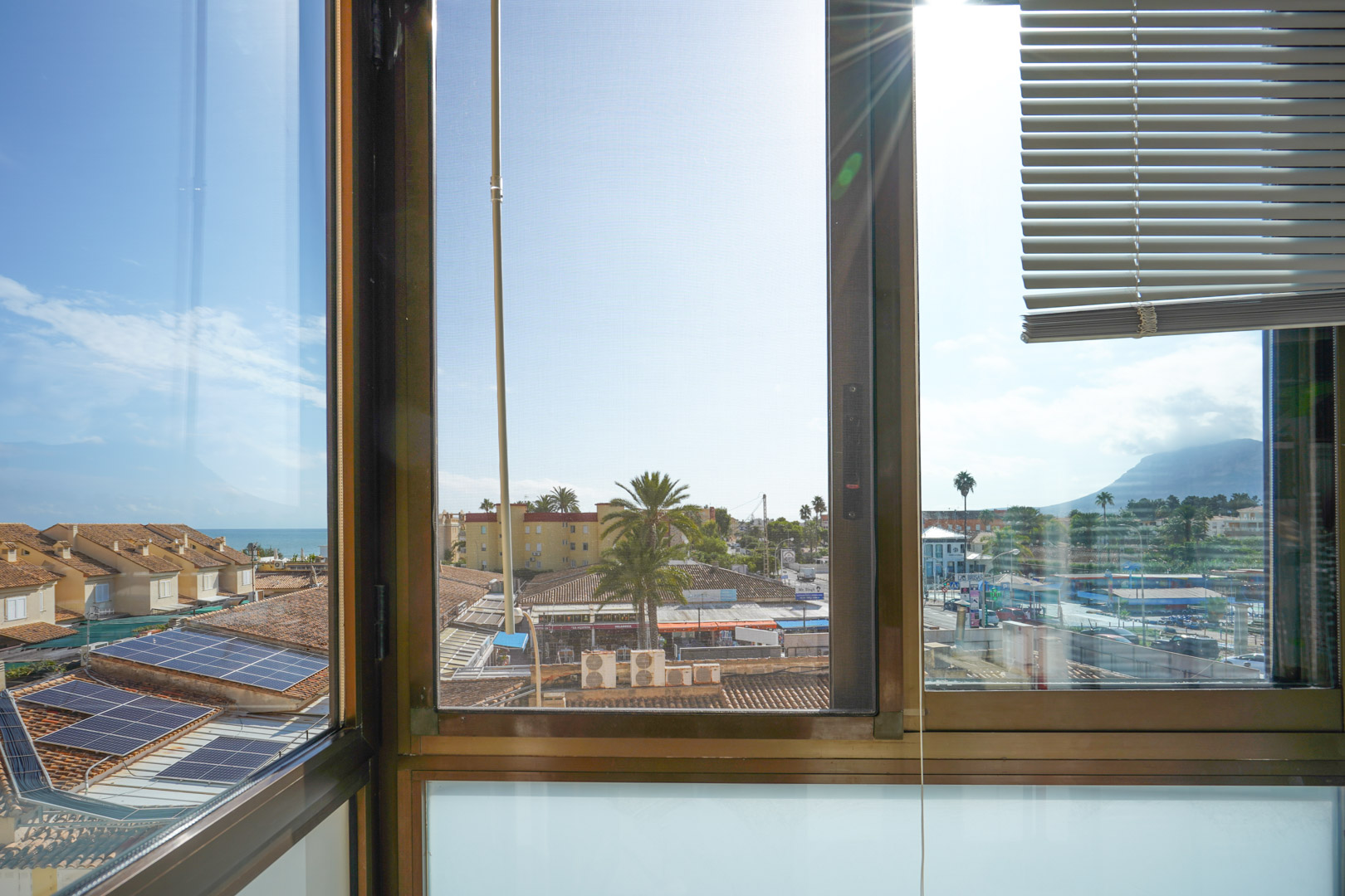 Blick durch ein Fenster mit Jalousien und Insektenschutz auf Ziegeldächer mit Solaranlagen, Palmen sowie das Meer und die Berge im Hintergrund.