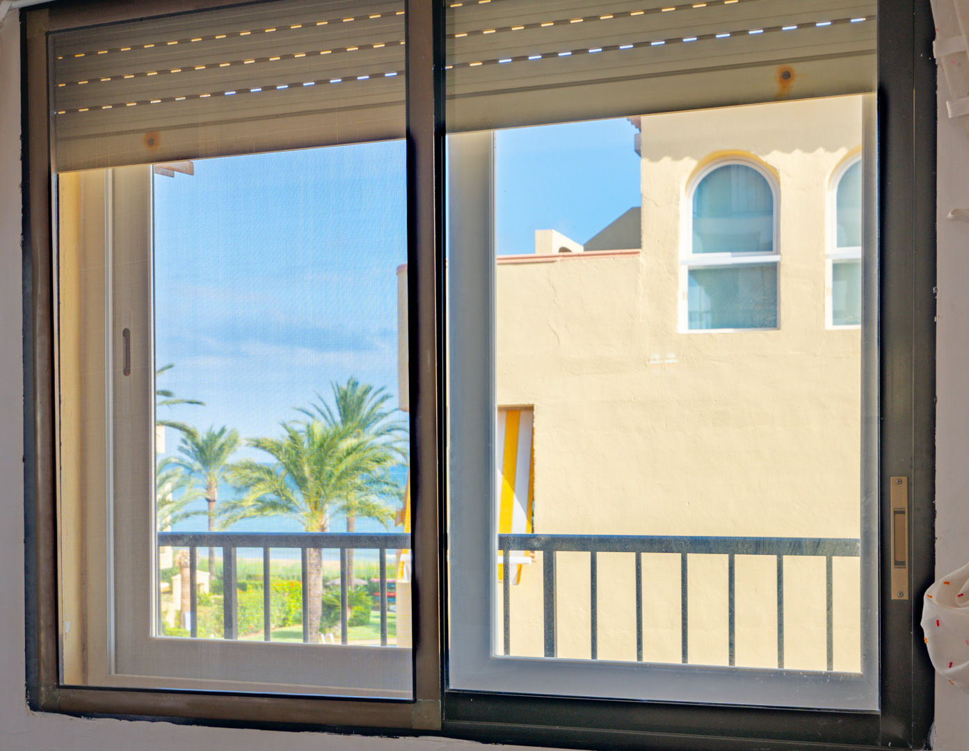Blick durch ein Schiebefenster mit Rollläden auf Palmen, das Meer im Hintergrund und mediterrane Architektur unter einem klaren blauen Himmel.