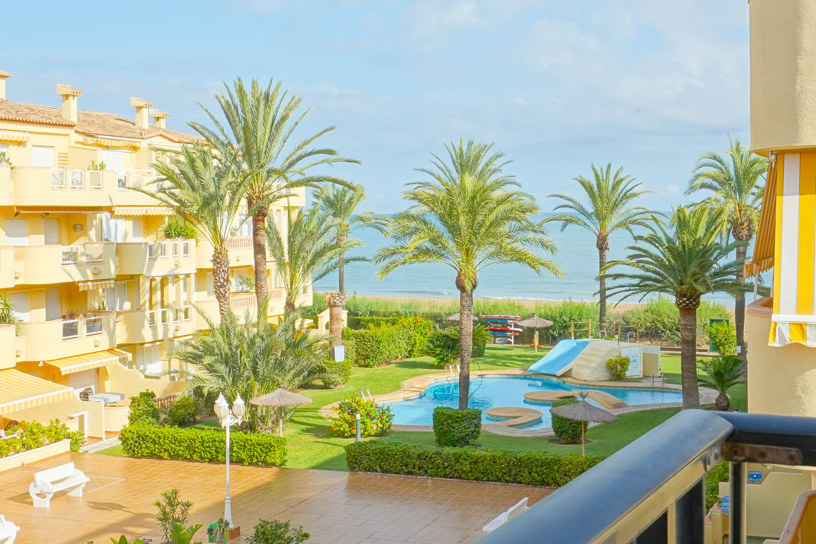 Blick vom privaten Balkon auf die gepflegte Gartenanlage mit Palmen, Gemeinschaftspool mit Rutsche und direktem Blick auf das Mittelmeer.