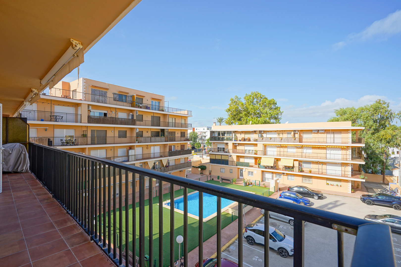 Blick von einem privaten Balkon mit Fliesenboden und Metallgeländer auf den Gemeinschaftspool, den Kunstrasen und den Parkplatz der Wohnanlage.