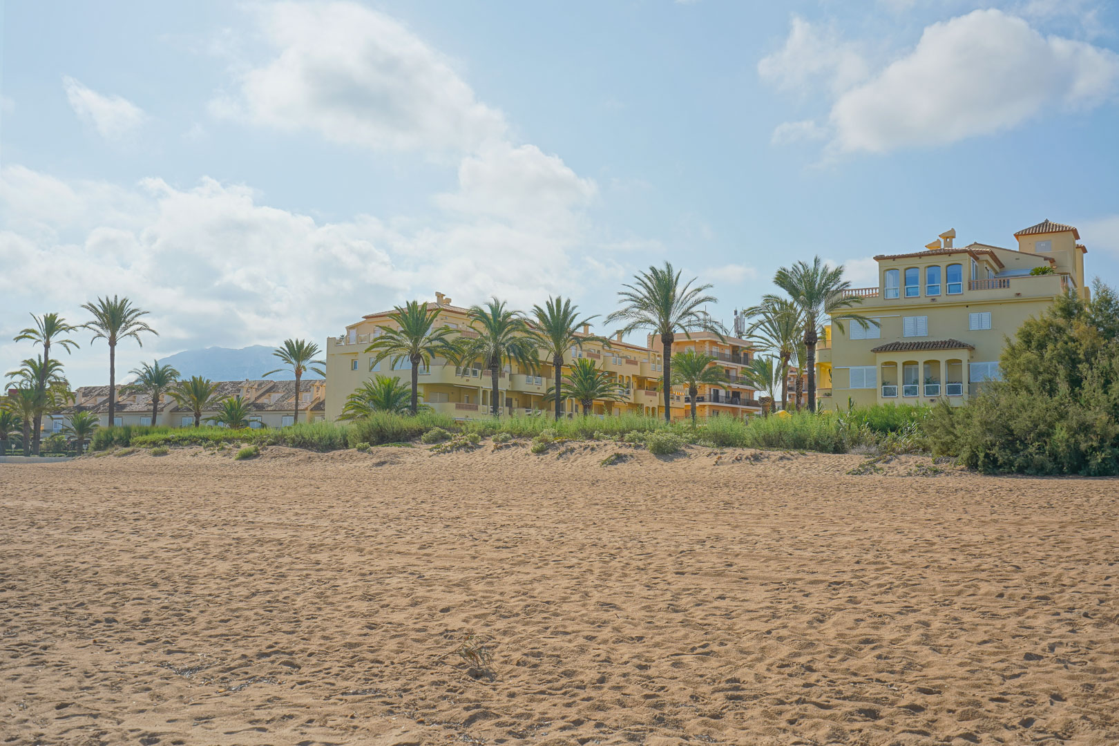 Breiter Sandstrand vor Wohngebäuden im mediterranen Stil mit Palmen und Blick auf die fernen Berge unter hellem Himmel.