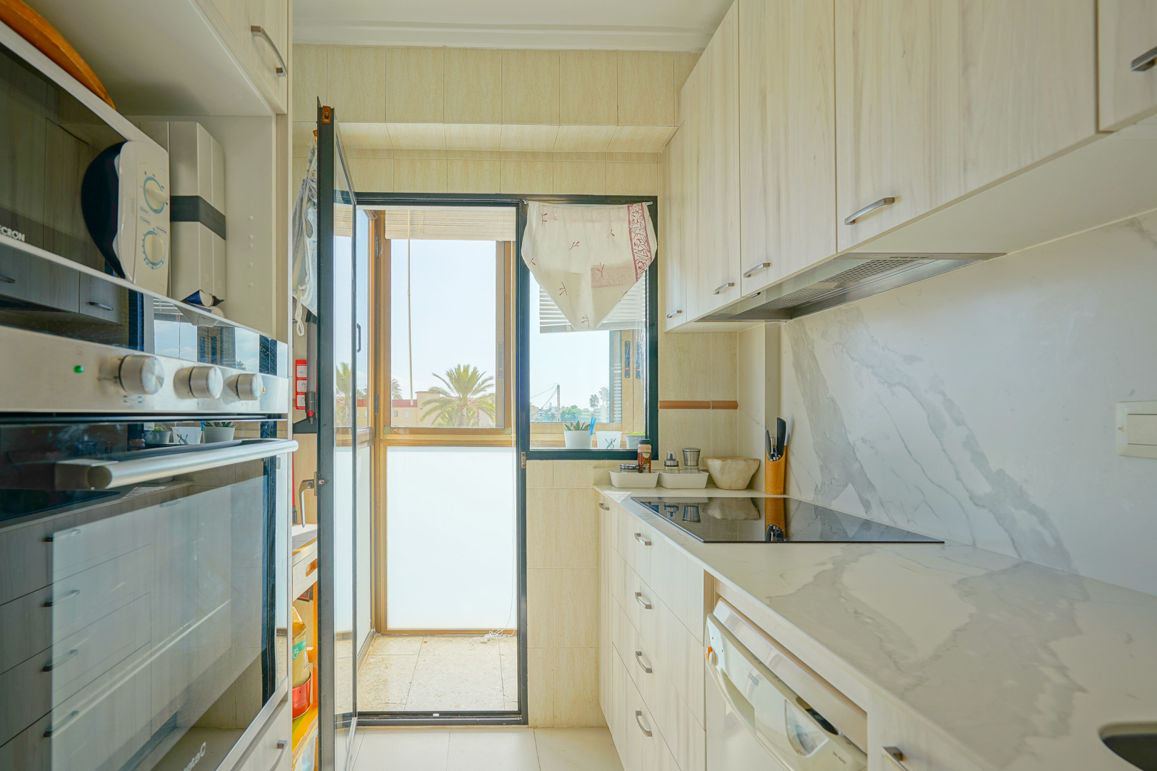 Einbauküche mit hellen Holzfronten, Arbeitsplatte in Marmoroptik und Glastür zu einem Balkon mit Blick auf Palmen.