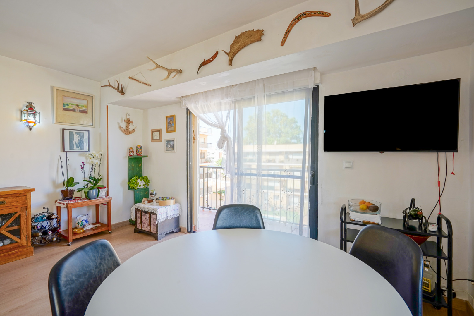 Essbereich mit rundem weißem Tisch, Wand-TV und Zugang zum Balkon. Einzigartige Wanddekoration mit Geweihen und Bumerangs ziert den Balken über dem Fenster.