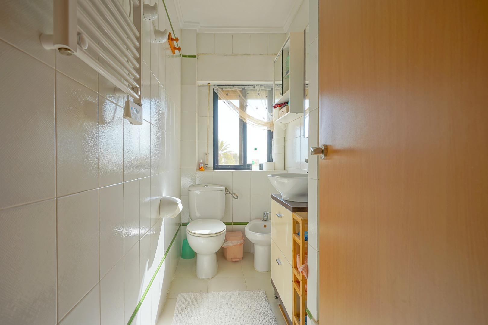 Funktionales Badezimmer mit weißen Fliesen, Bidet, WC und Waschbecken. Ein Fenster sorgt für natürliches Licht und bietet einen Blick auf die grüne Umgebung.