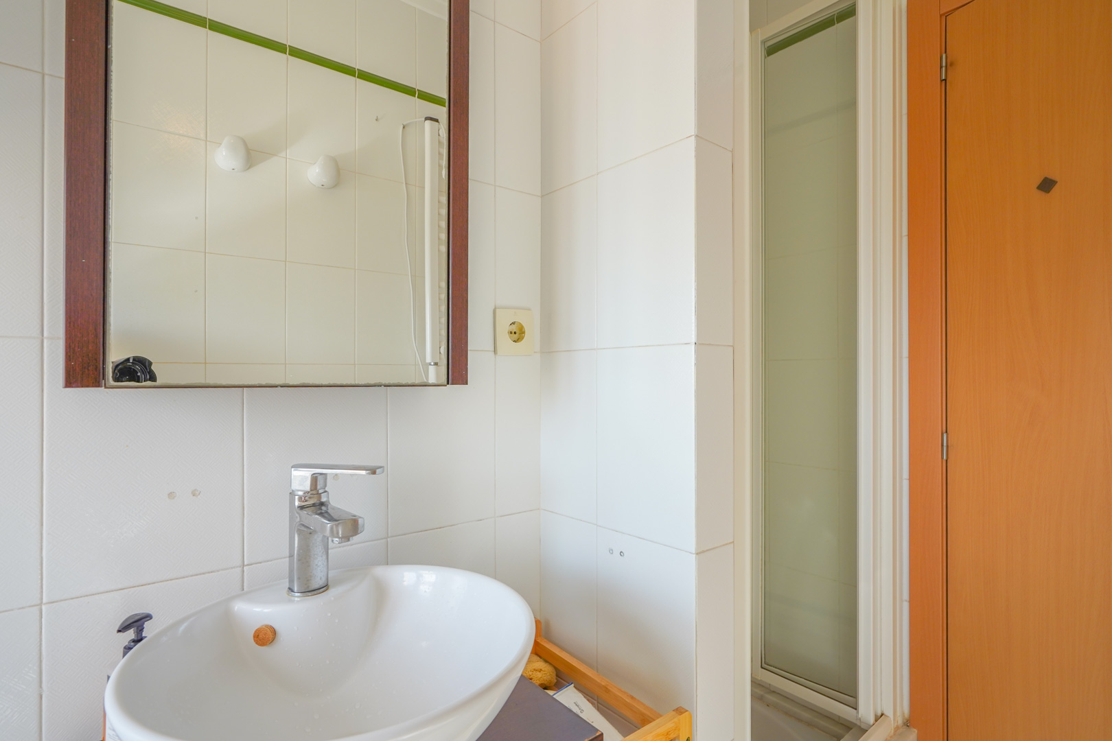 Funktionelles Badezimmer mit weißem Keramikwaschbecken, verchromter Armatur und Holzrahmen-Spiegel neben einer Dusche mit mattierter Glastür.