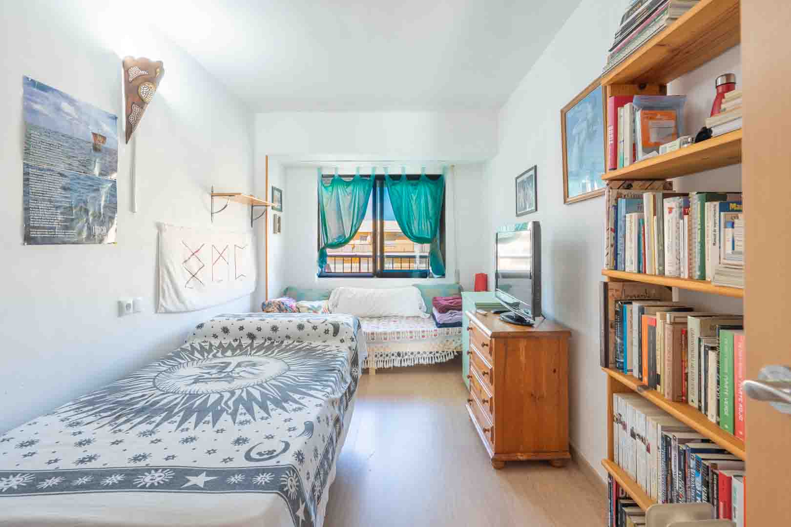 Helles Schlafzimmer mit Einzelbett, Holzregal und Kommode mit Fernseher. Natürliches Licht fällt durch ein Fenster mit türkisfarbenen Vorhängen ein.